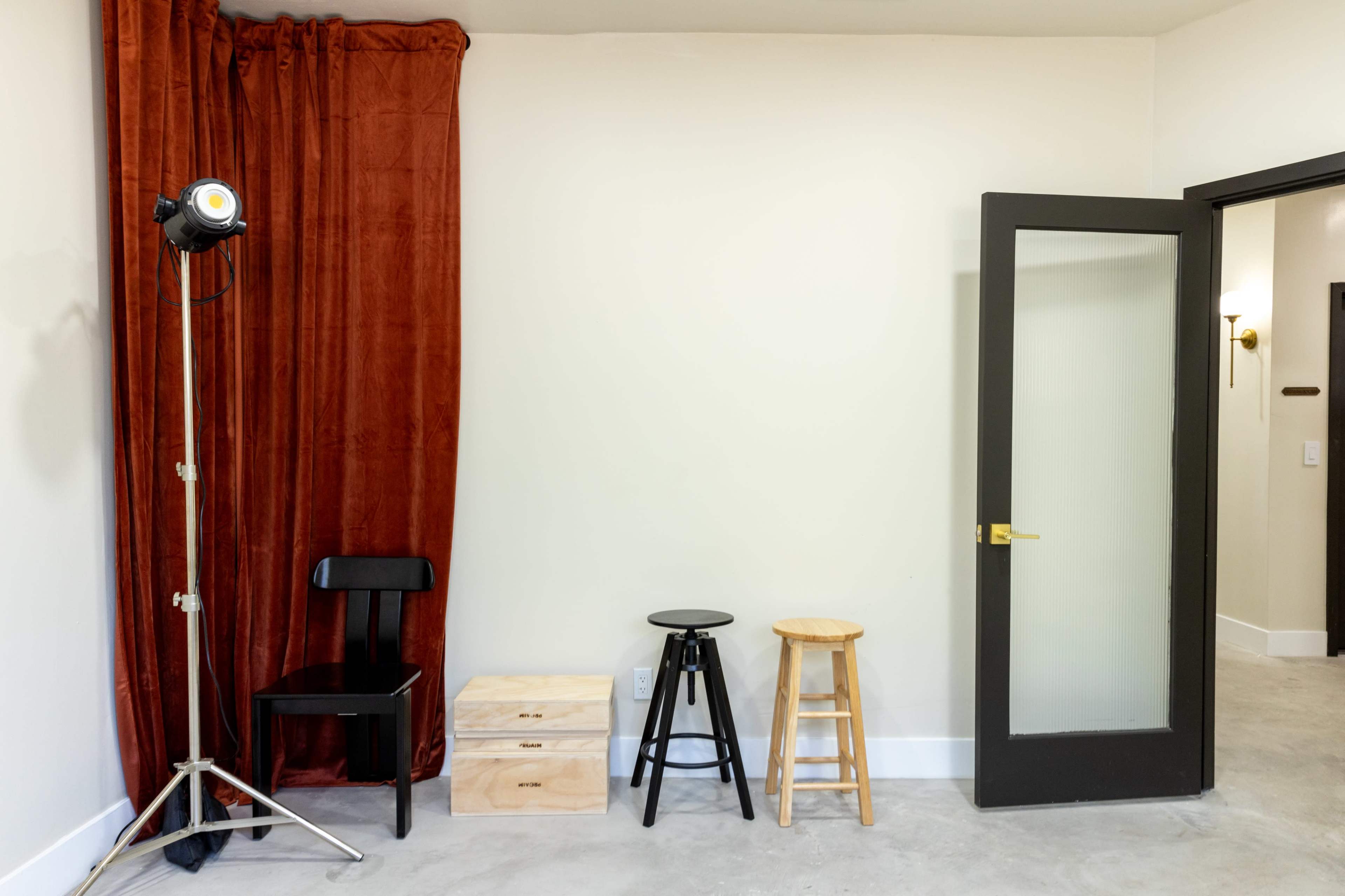 A small room with a red curtain, a spotlight on a stand, and three stools positioned against a neutral wall, along with an open door.