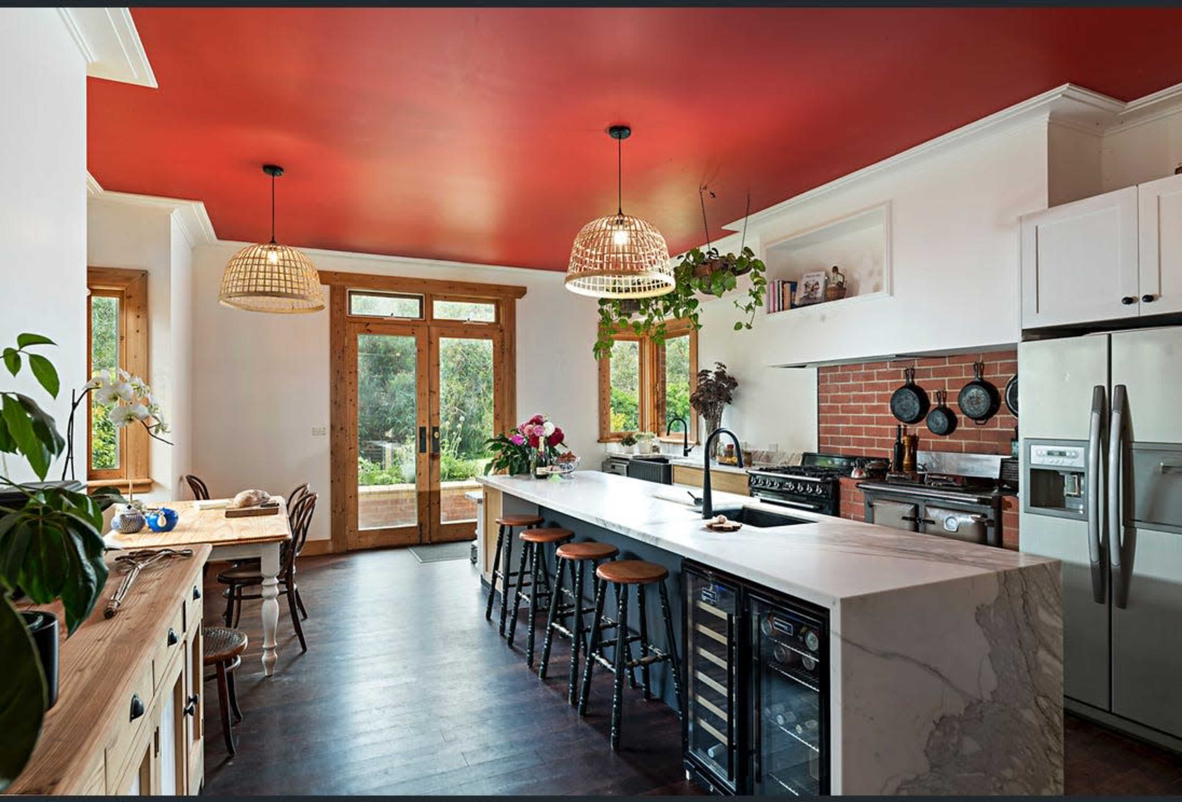 A modern kitchen features a large marble island, wooden cabinetry, pendant lighting, and a red ceiling, with large windows offering views of the outdoors.