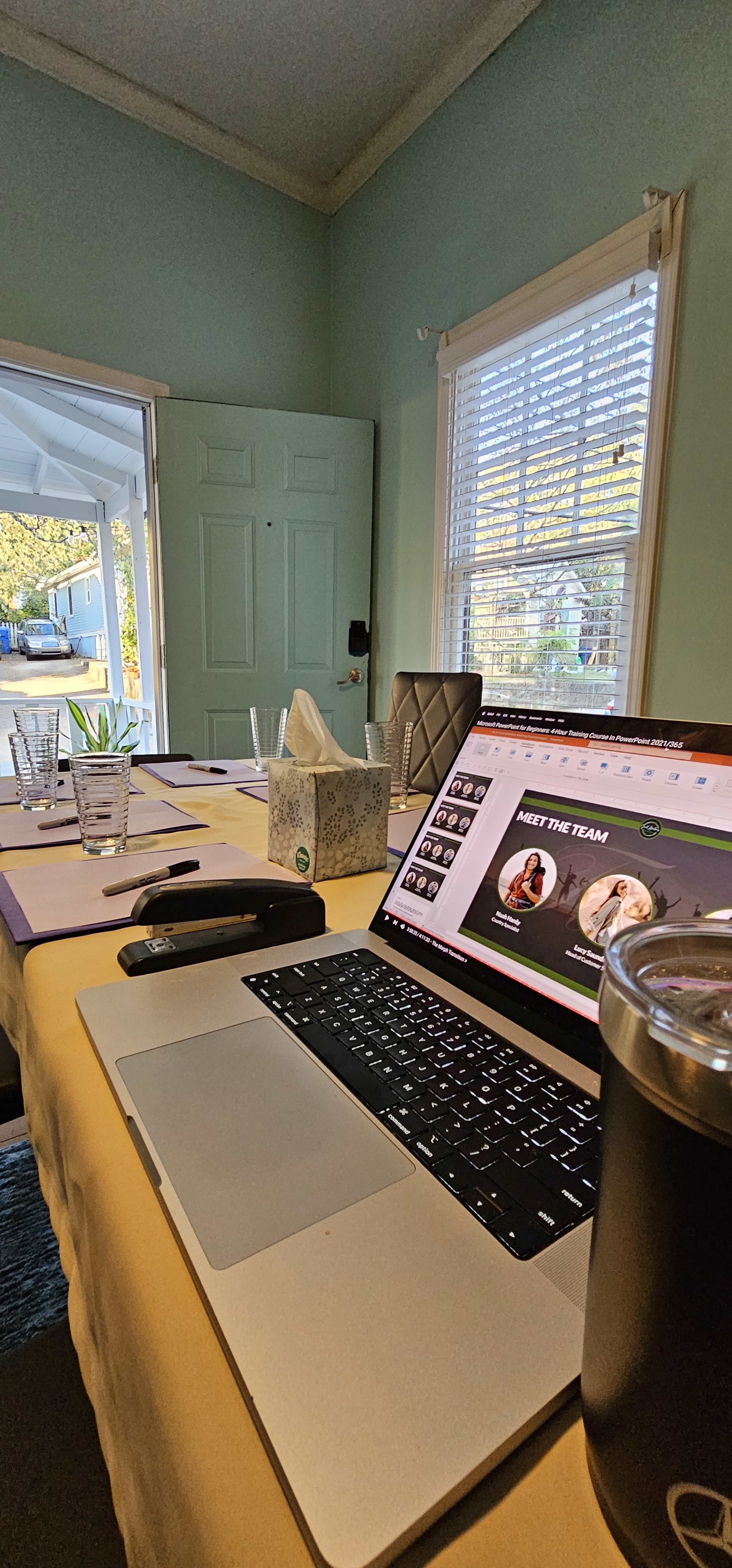 A laptop displaying a presentation is placed on a table near a tissue box and a drink, with a sunlit doorway visible in the background.
