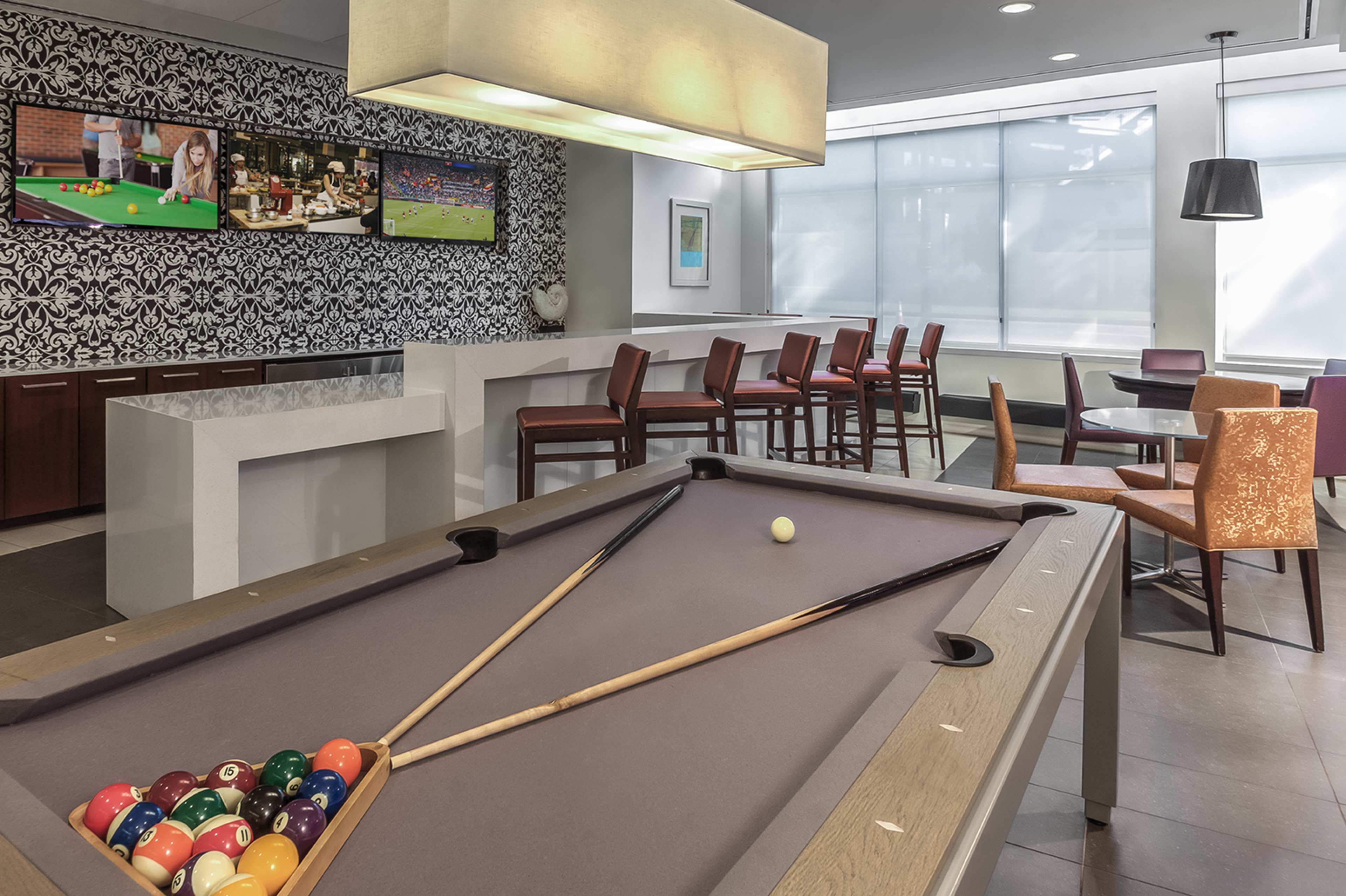 A modern lounge area featuring a pool table with a triangle of balls, a bar with high stools, and a patterned wallpaper with TV screens displaying sports.
