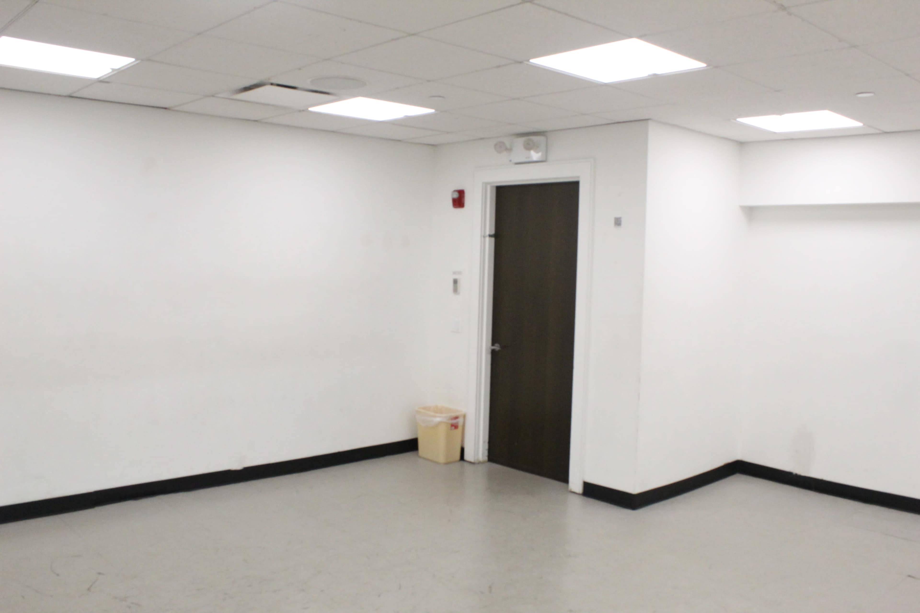 The image shows a sparsely furnished room with plain white walls, a dark door, and a small trash bin in the corner.