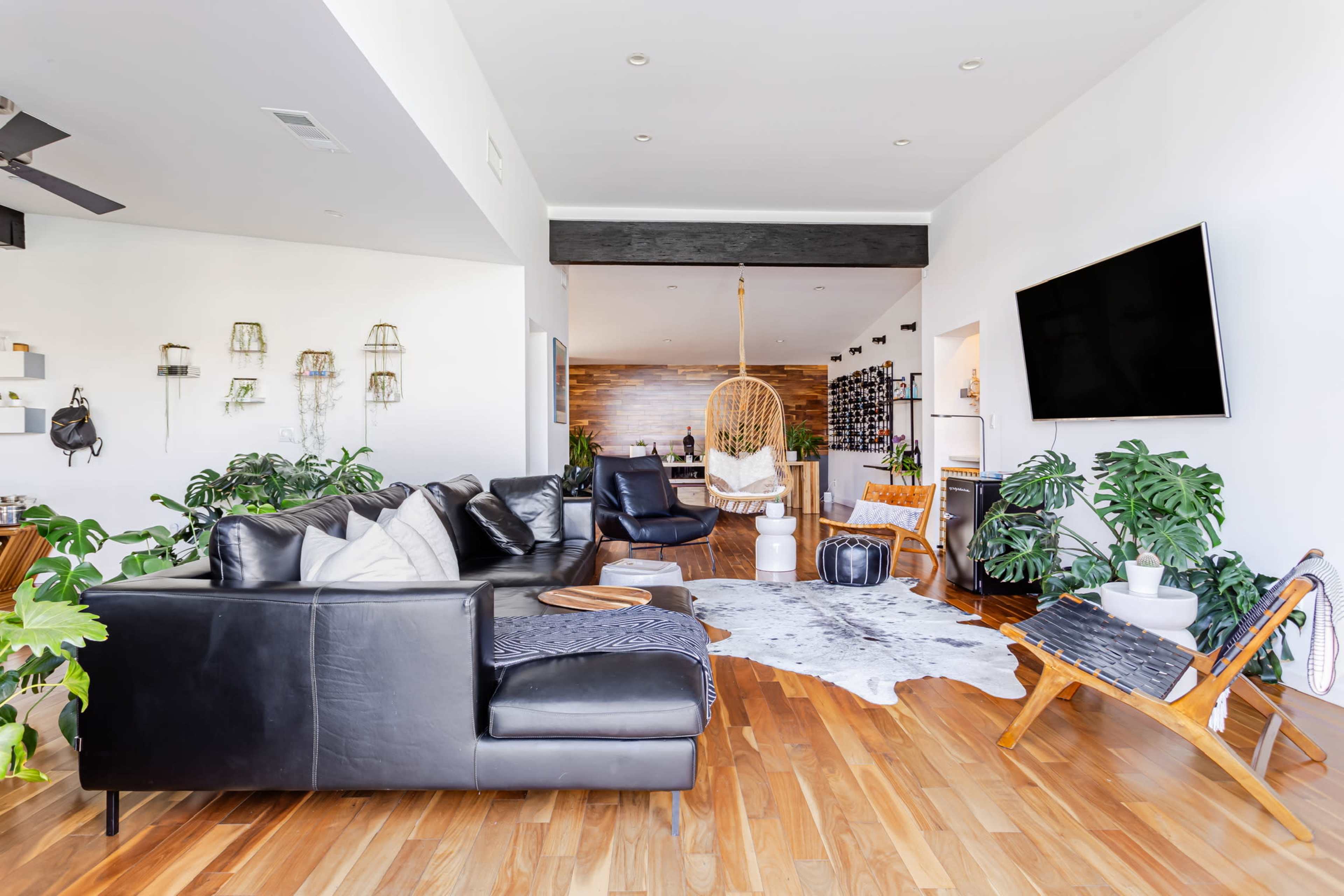 The image shows a modern living room with a black leather sectional couch, wooden flooring, and various indoor plants, featuring a hanging chair in the background.