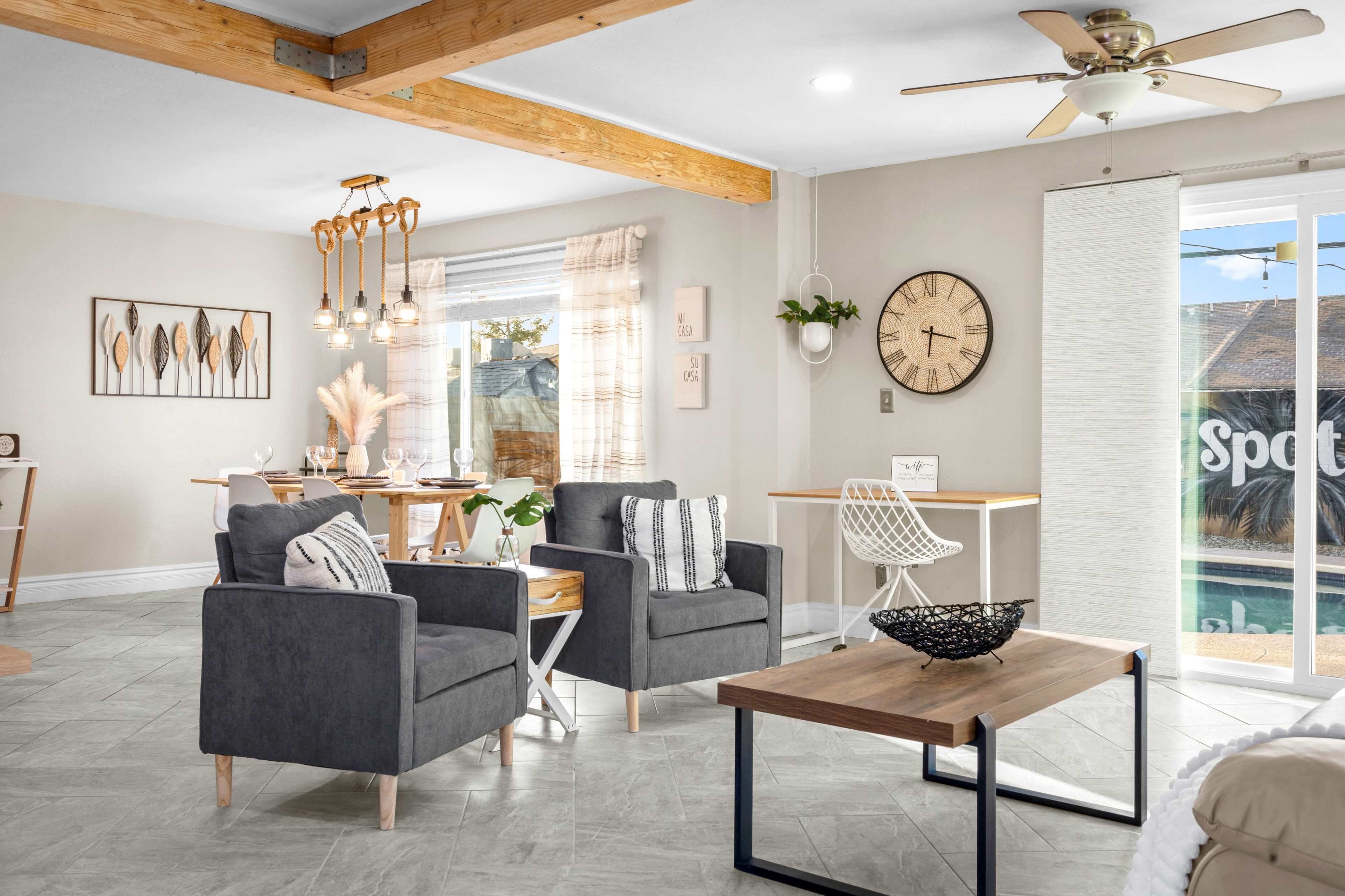A modern living room features two gray armchairs, a wooden coffee table, and a dining area with a light fixture and multiple chairs.