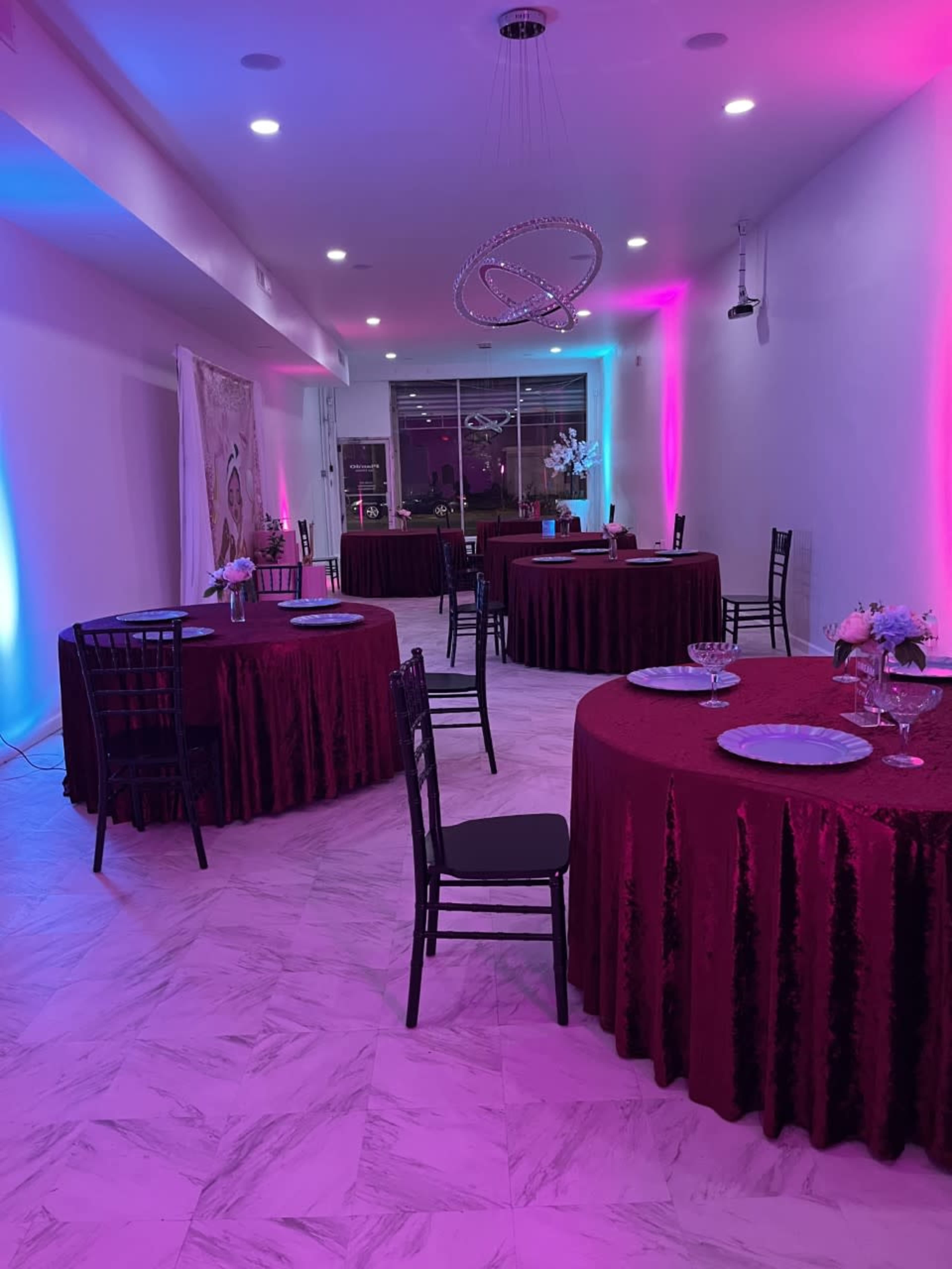A banquet hall set up with several round tables covered in maroon tablecloths, surrounded by black chairs, and illuminated by ambient lighting in pink and blue hues.