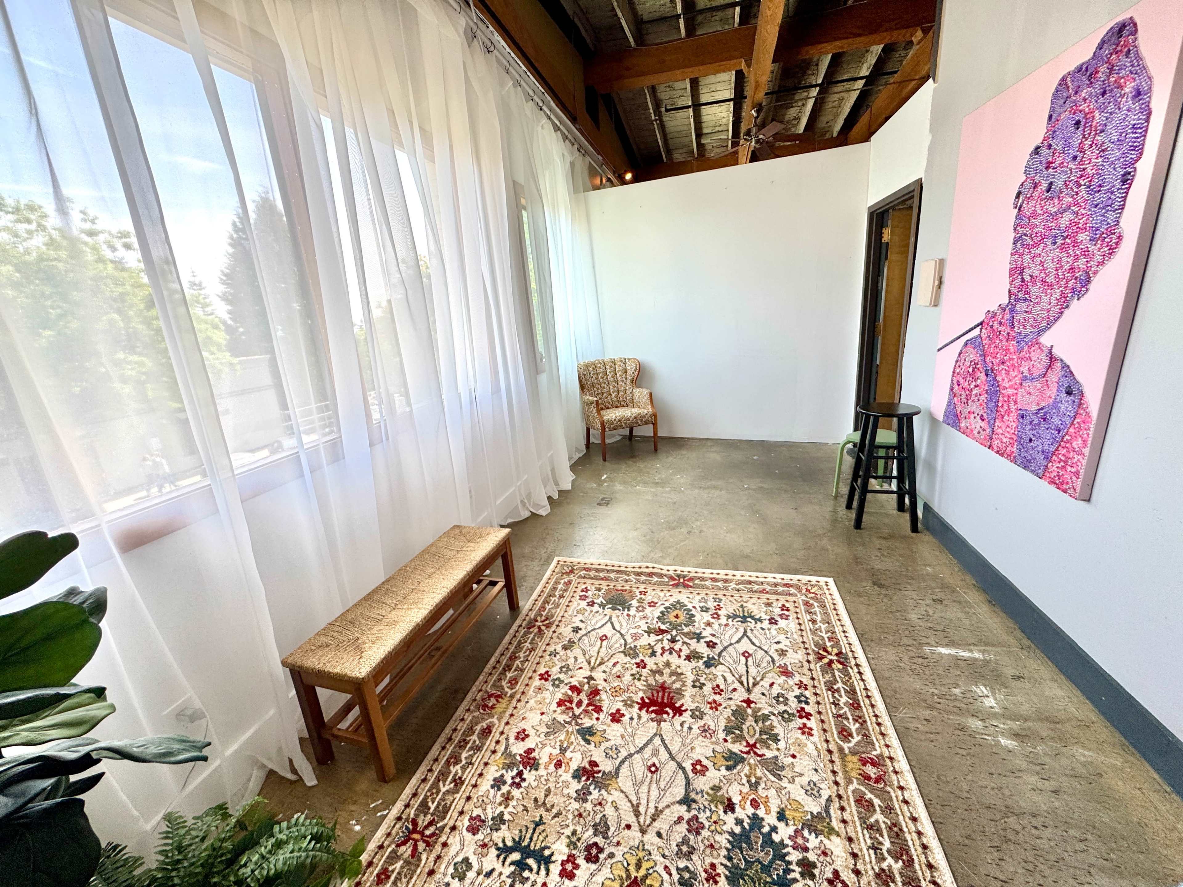 The image shows a bright, airy room with sheer curtains, a patterned rug on the concrete floor, a wooden bench, a chair, and a large artwork on the wall.