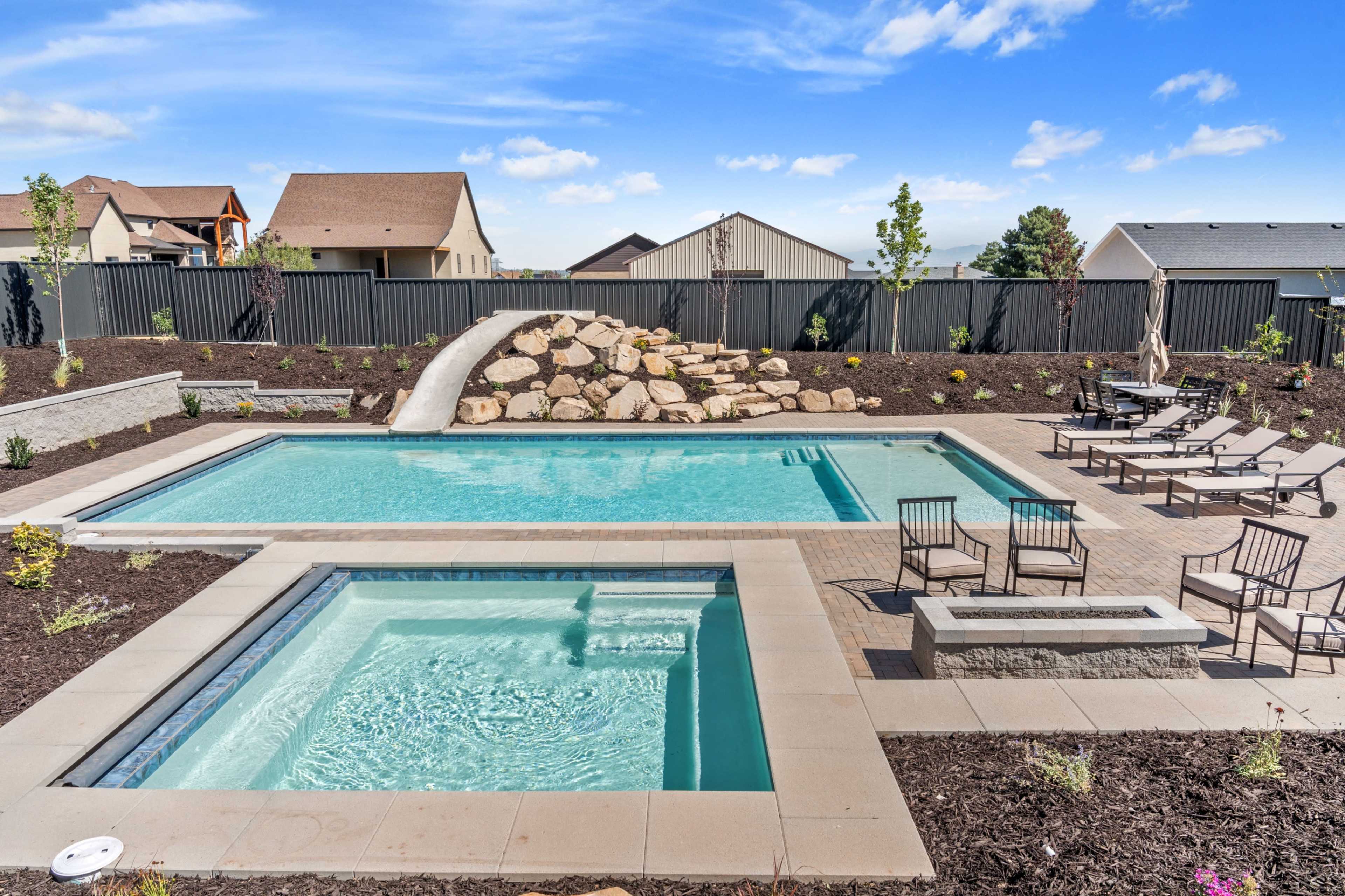 The image shows a landscaped backyard featuring a rectangular swimming pool, a spa area, and multiple seating areas with chairs along the pool's edge, surrounded by shrubs and decorative rocks.