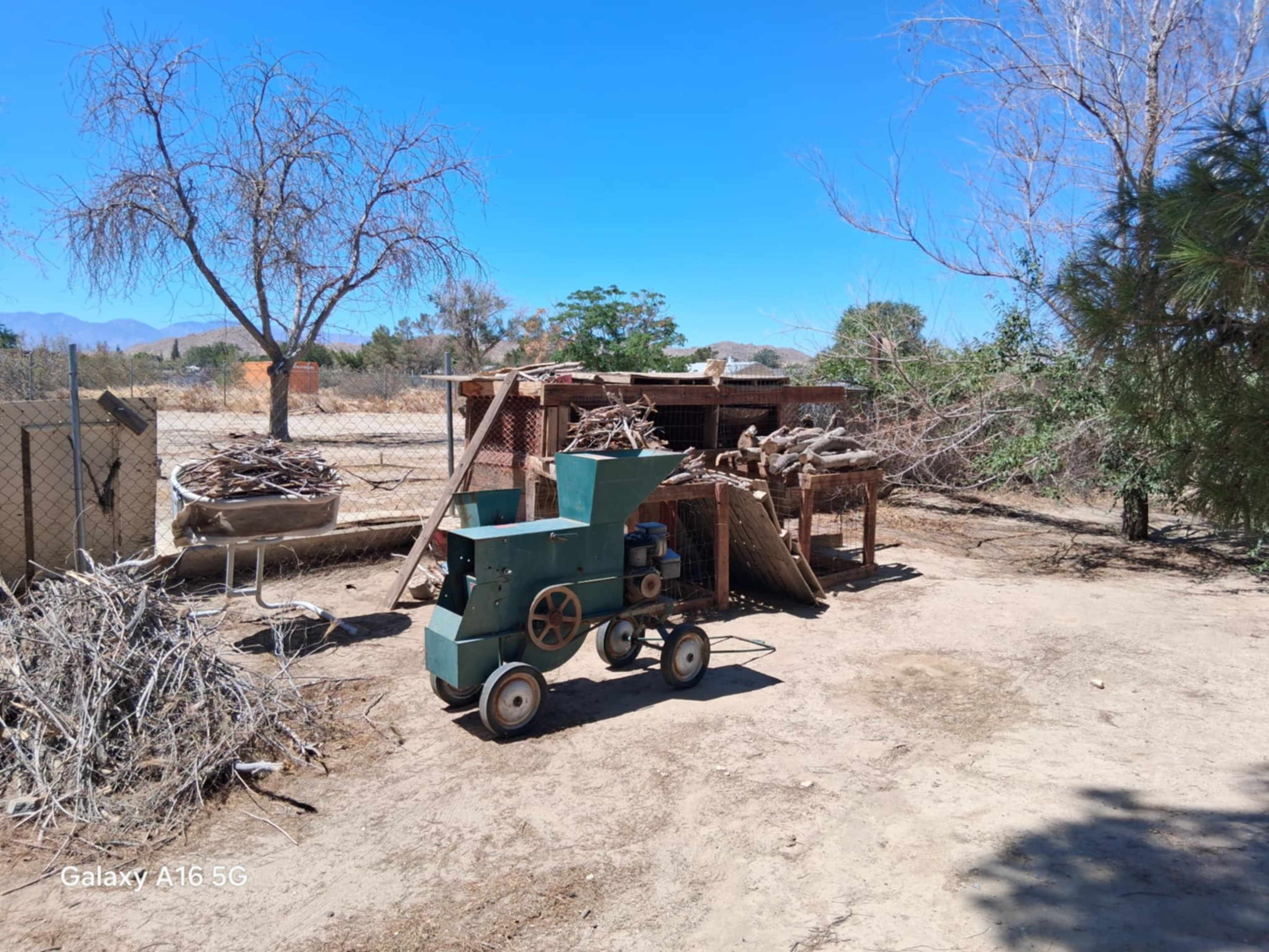 A small, vintage machine with wheels stands near a wooden shed surrounded by dry brush and trees in an open landscape.