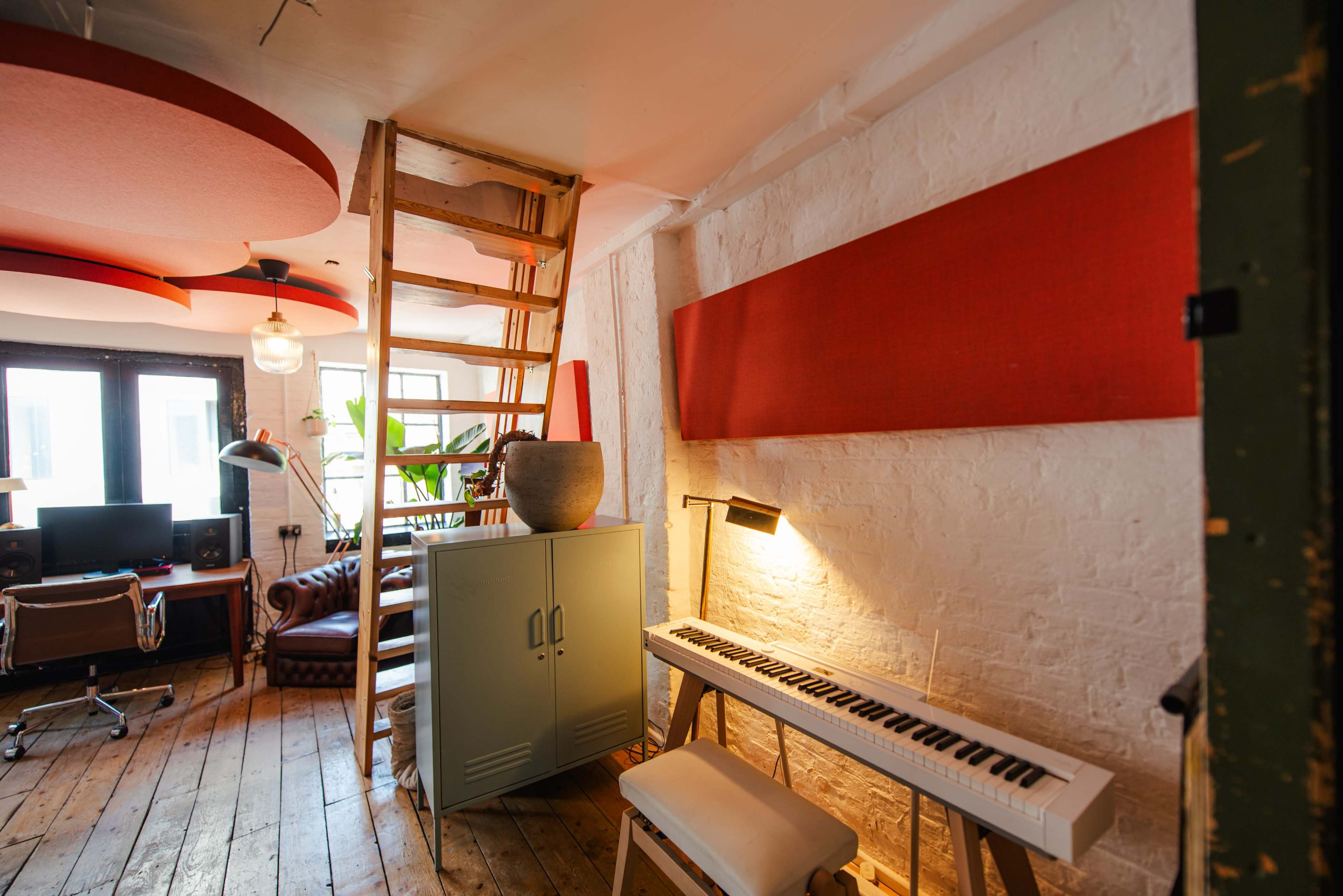 The image shows a stylish, modern workspace featuring a ladder leading to an upper level, a keyboard, and a plant beside a desk and seating area.