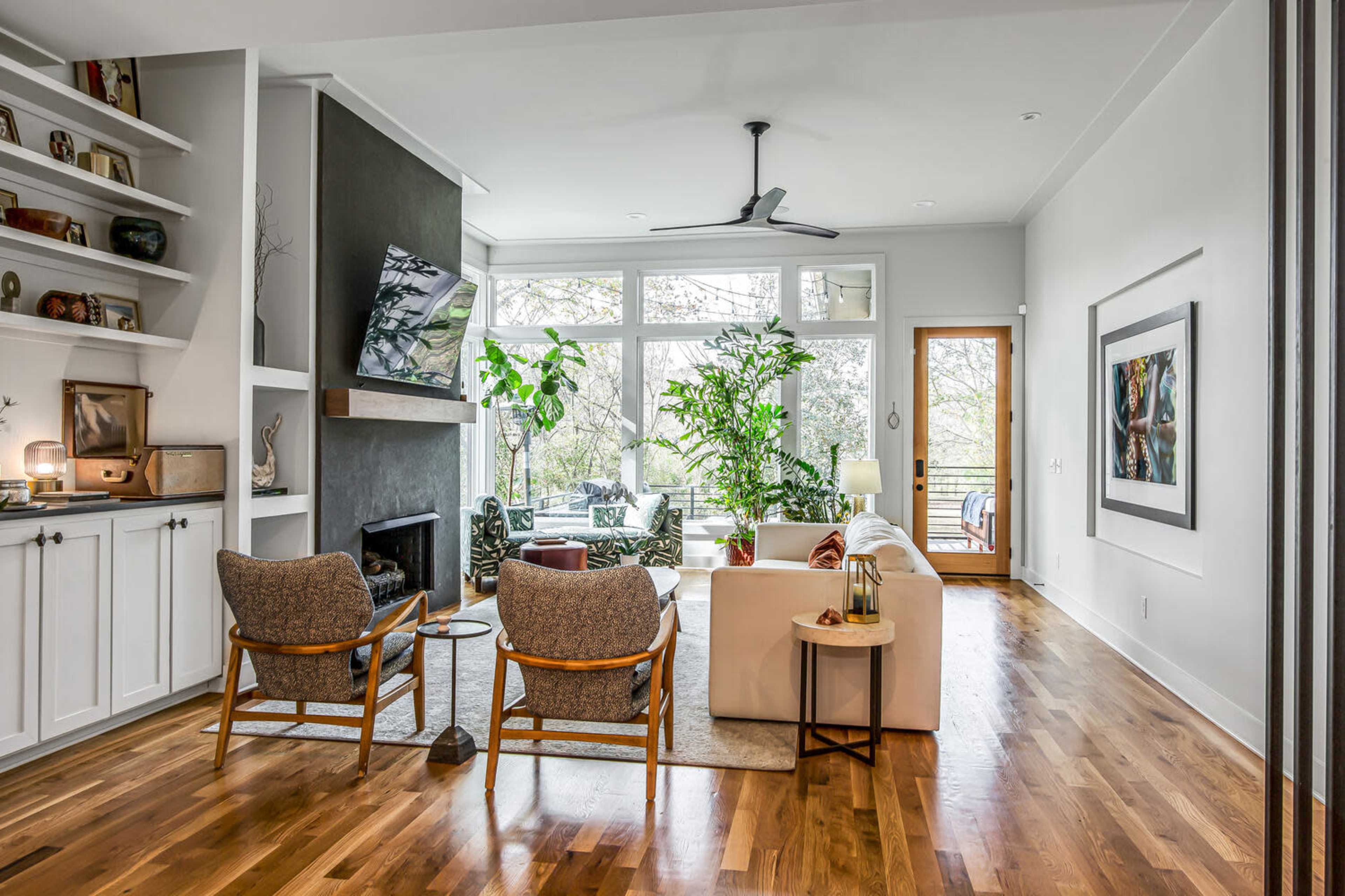 The image shows a modern living room with a large window, plants, a fireplace, and seating arranged around a coffee table.
