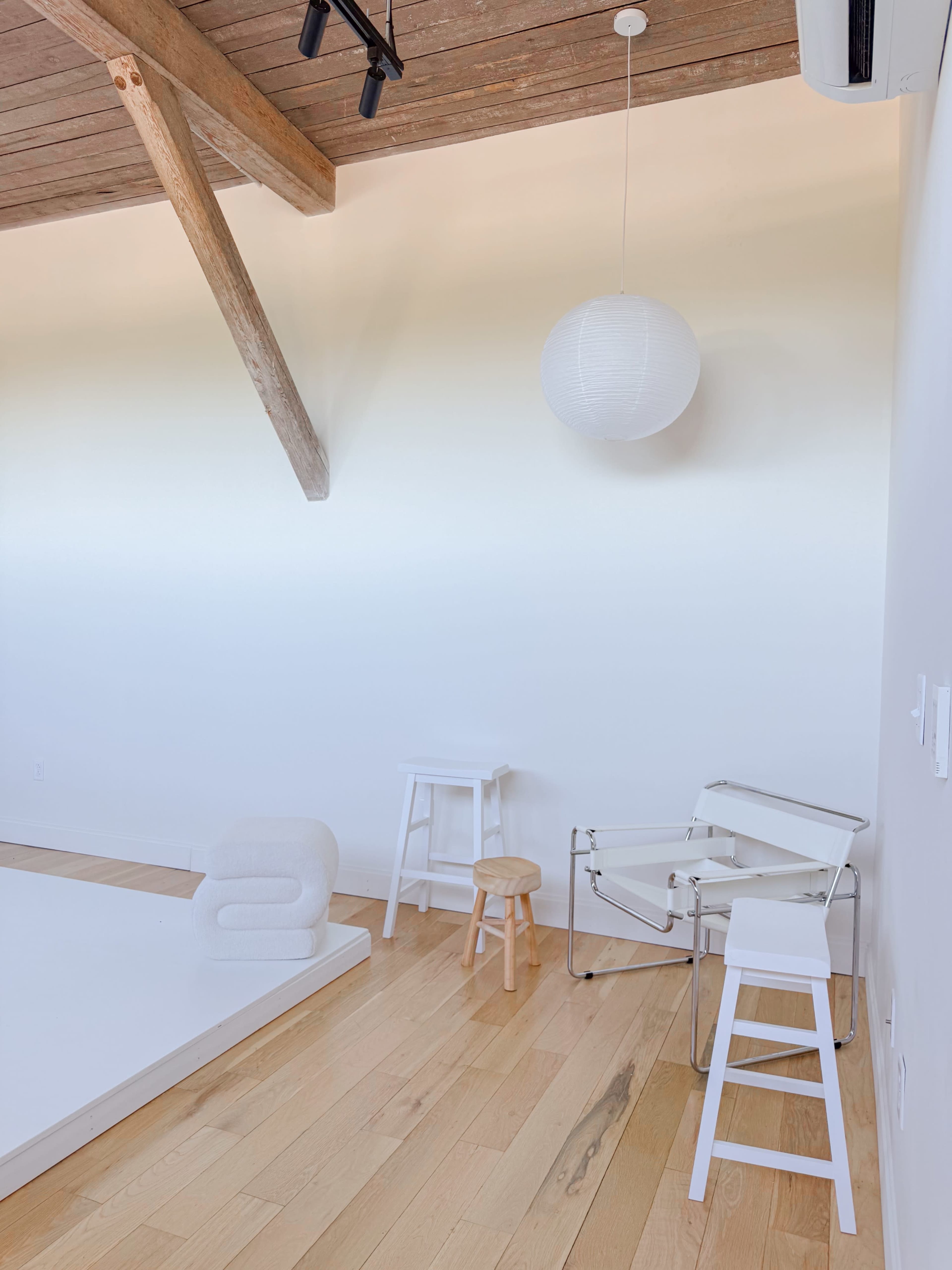 The image shows a minimalist interior space with wooden beams, a white ceiling, and various furniture pieces including stools and a chair.