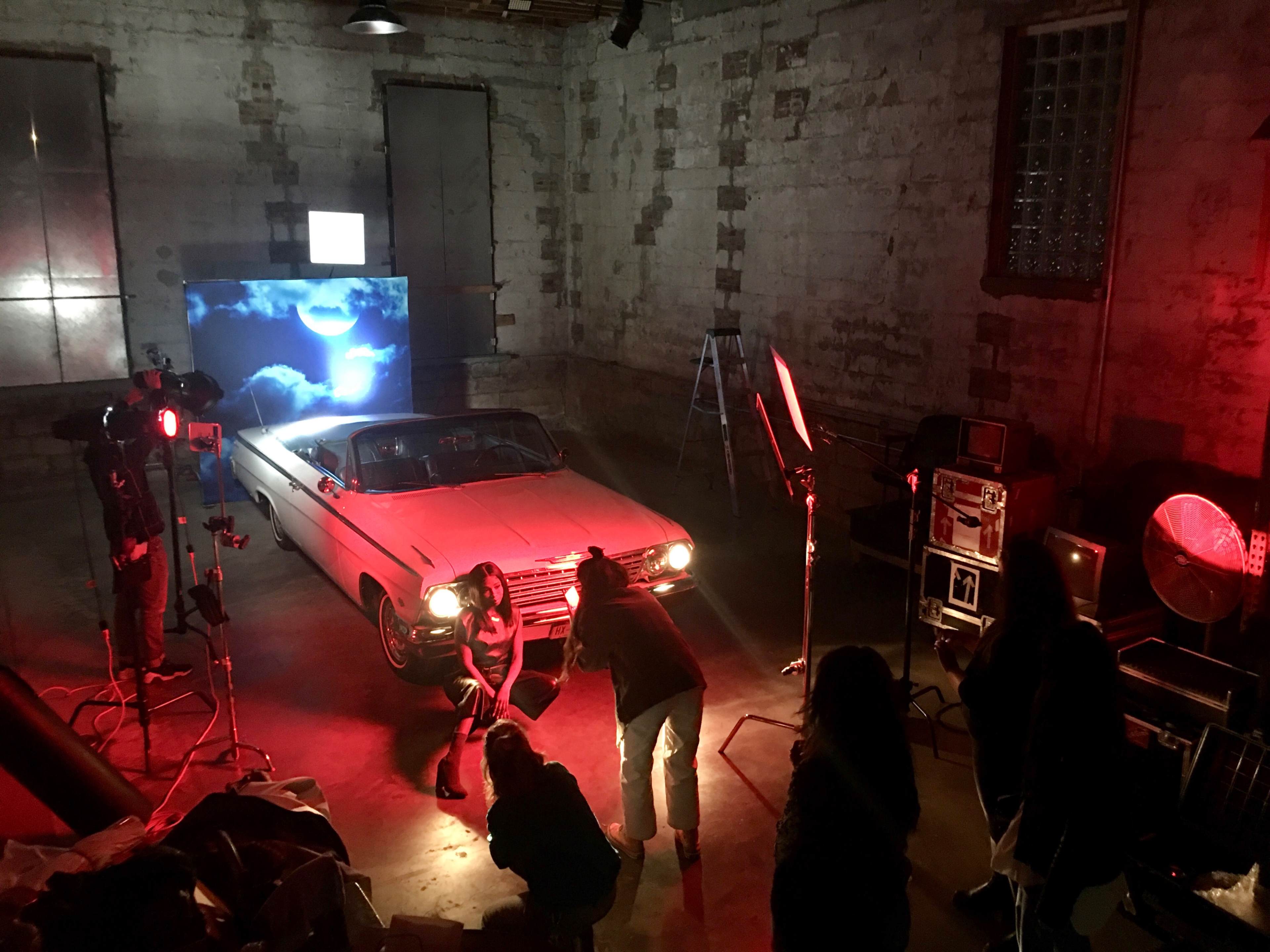 A film crew sets up lighting and equipment around a classic car in a dimly lit industrial space.