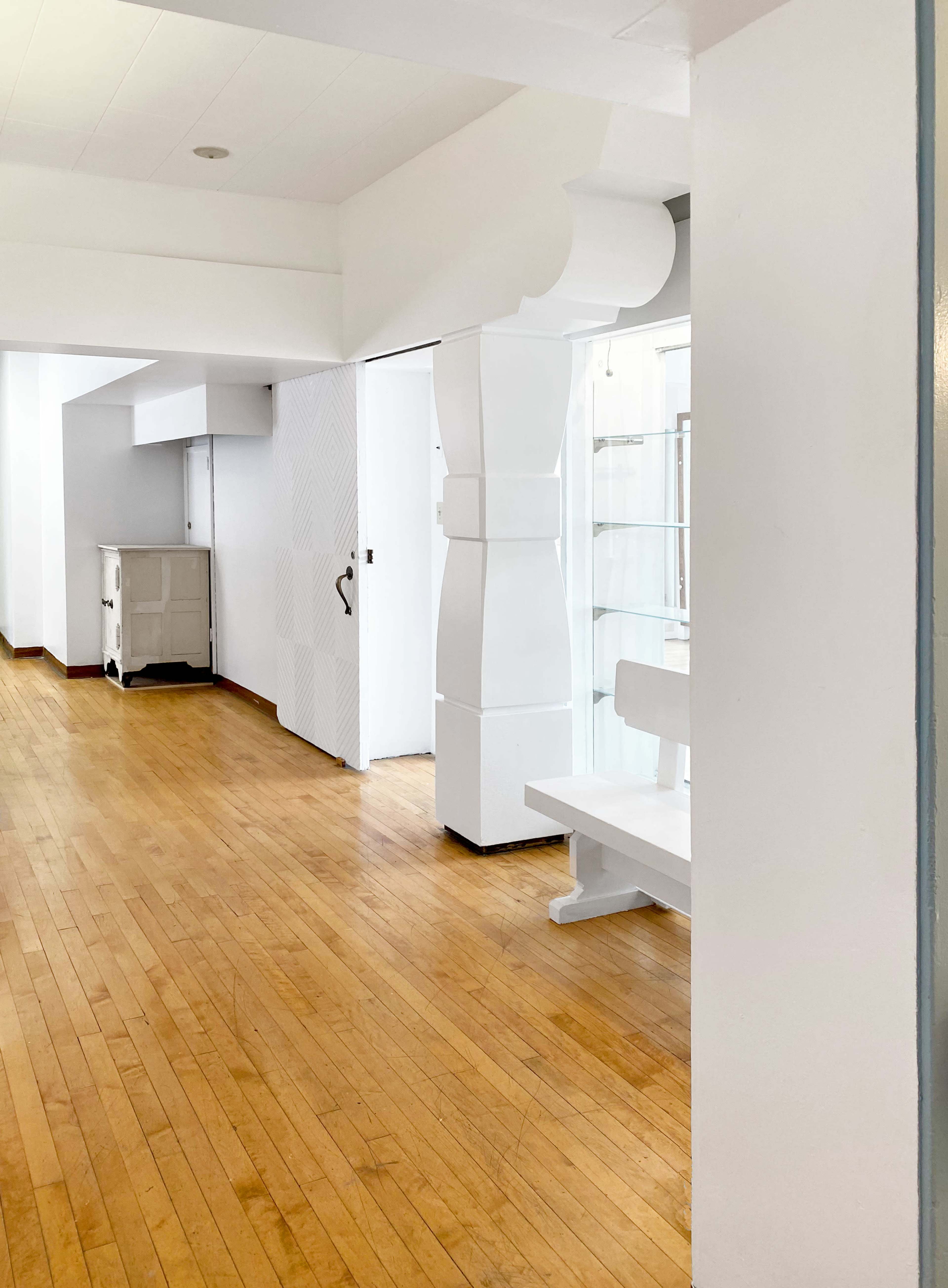 The image shows a bright, minimalist interior room with wooden floors, white walls, and a bench along one side.