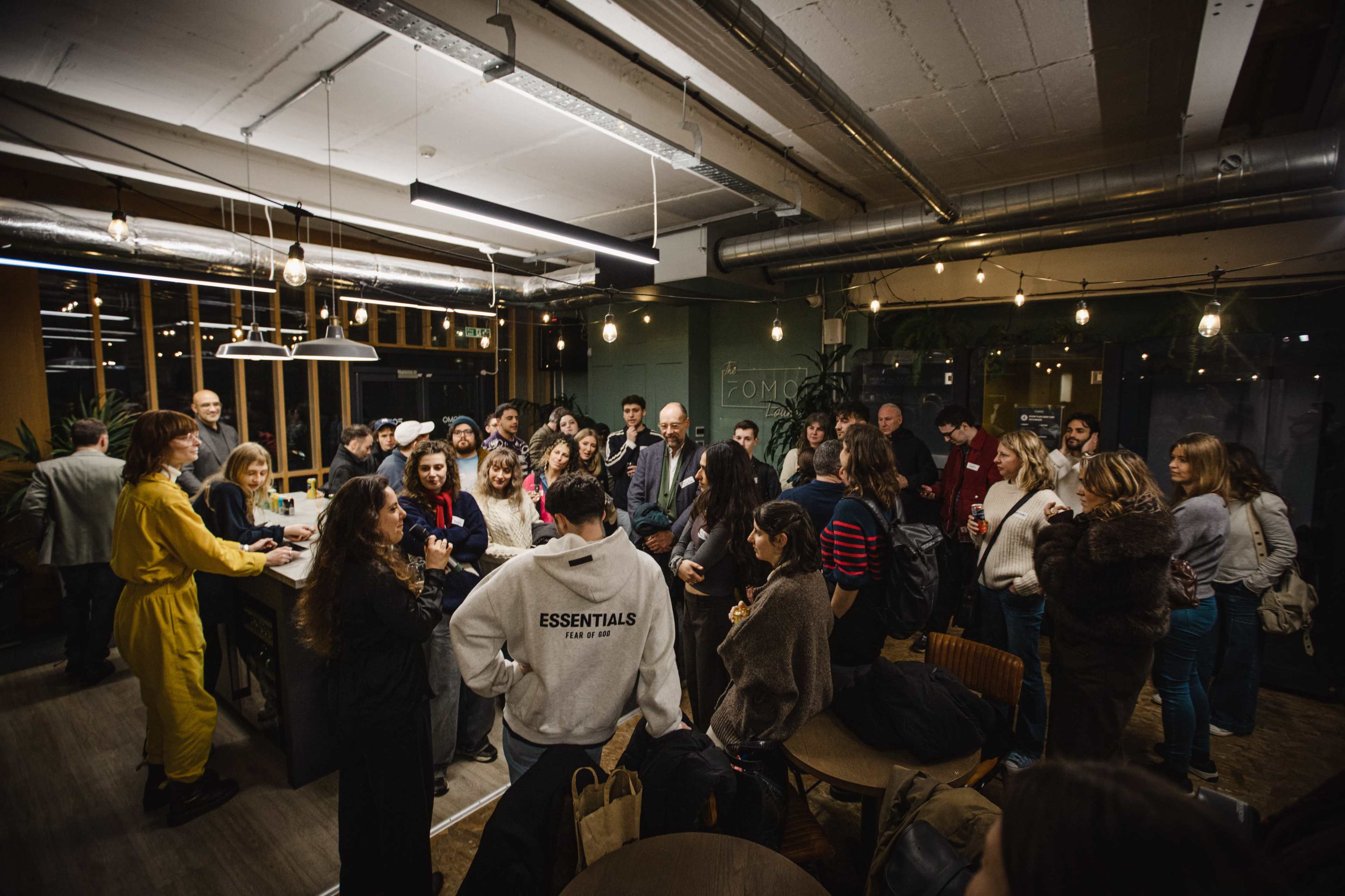 A large group of people gathers in a well-lit, modern room with plants and industrial decor, engaging in conversation around a central table.