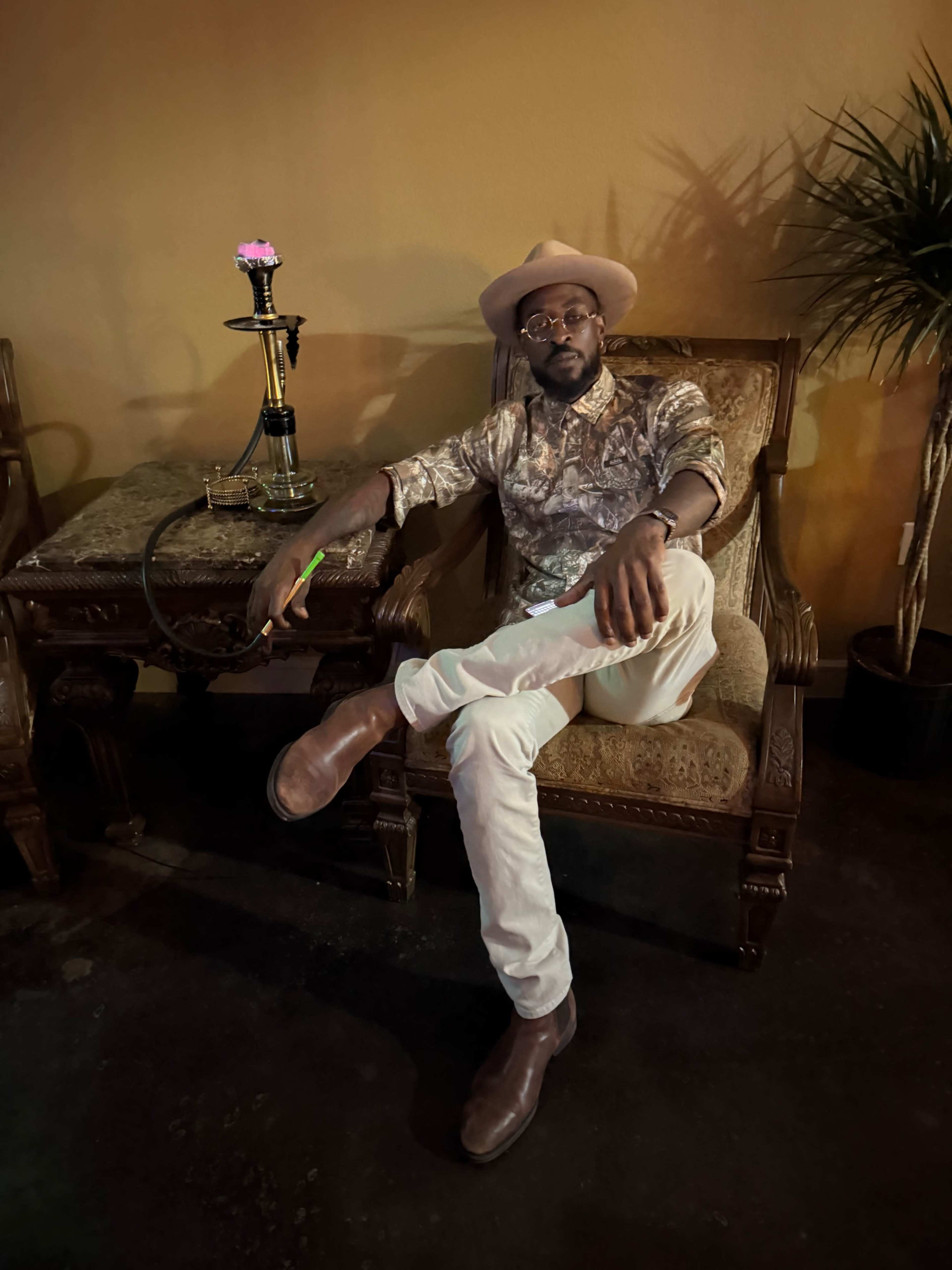 A man sits comfortably in a vintage chair, wearing a patterned shirt and hat, with a hookah beside him and a potted plant in the background.
