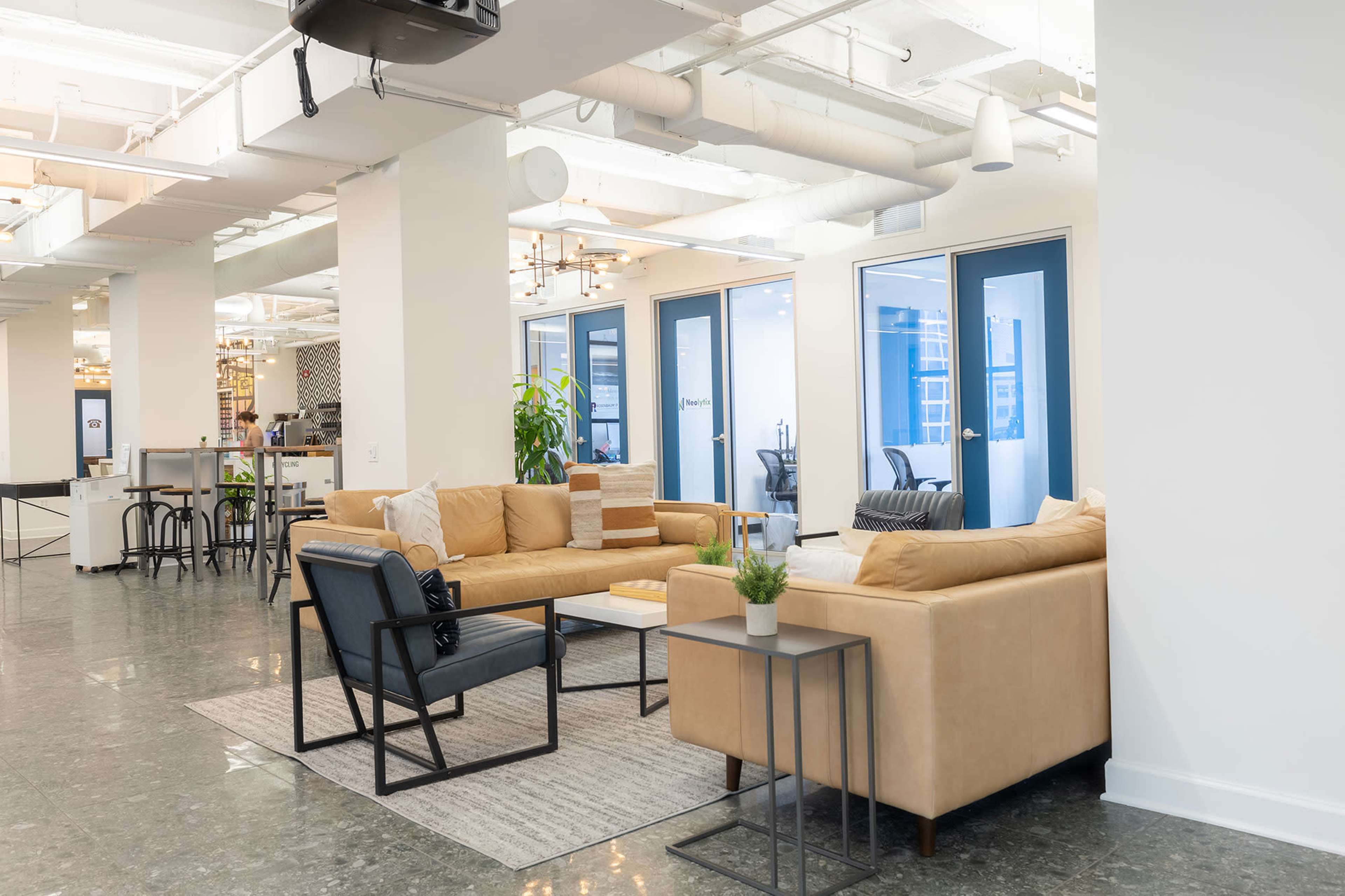 The image shows a modern office lounge area featuring a light brown sectional sofa, a black armchair, and a small side table on a patterned rug, with glass doors leading to other office spaces.