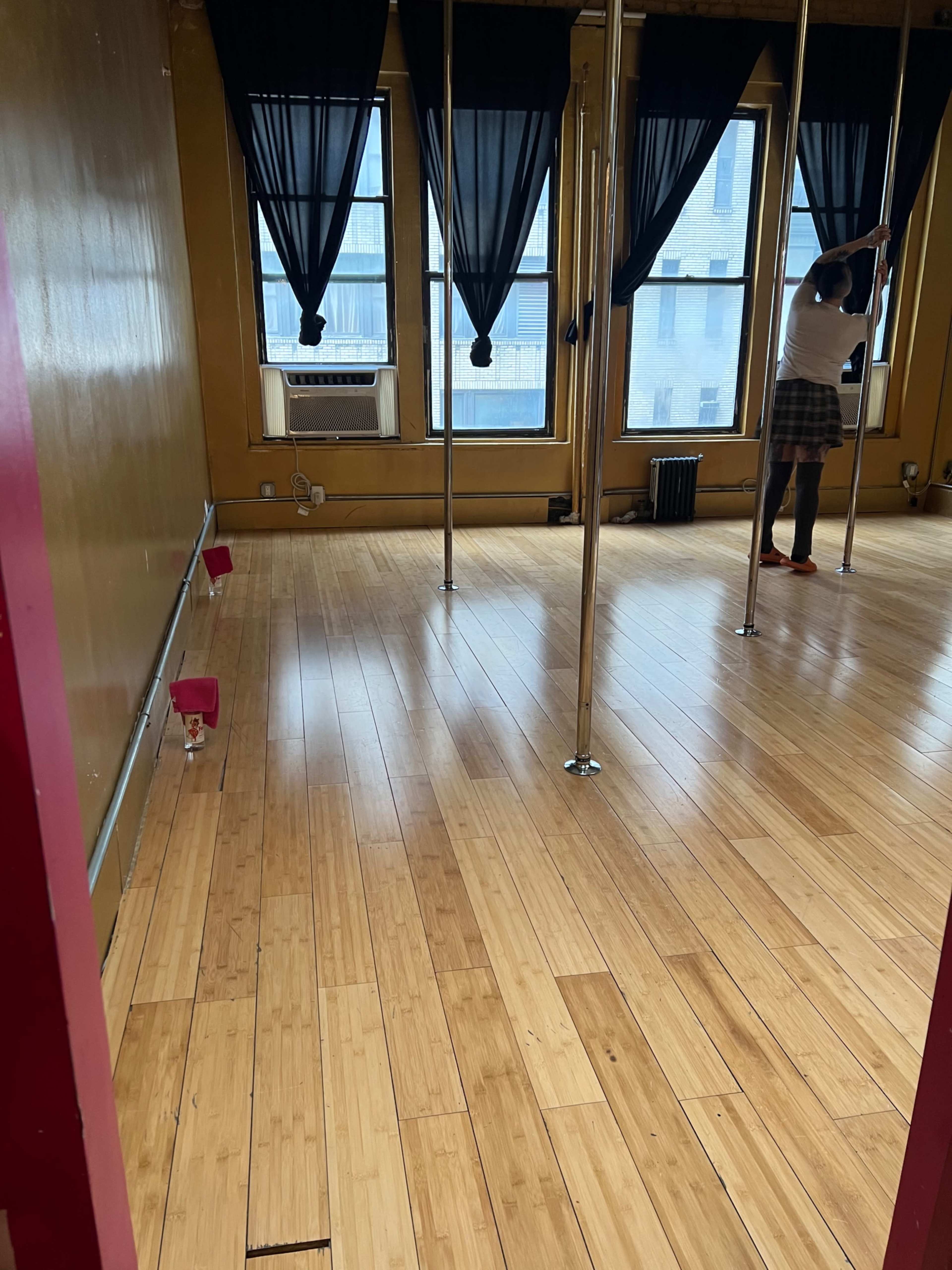 A room with bamboo flooring features several vertical poles and large windows draped with black curtains.