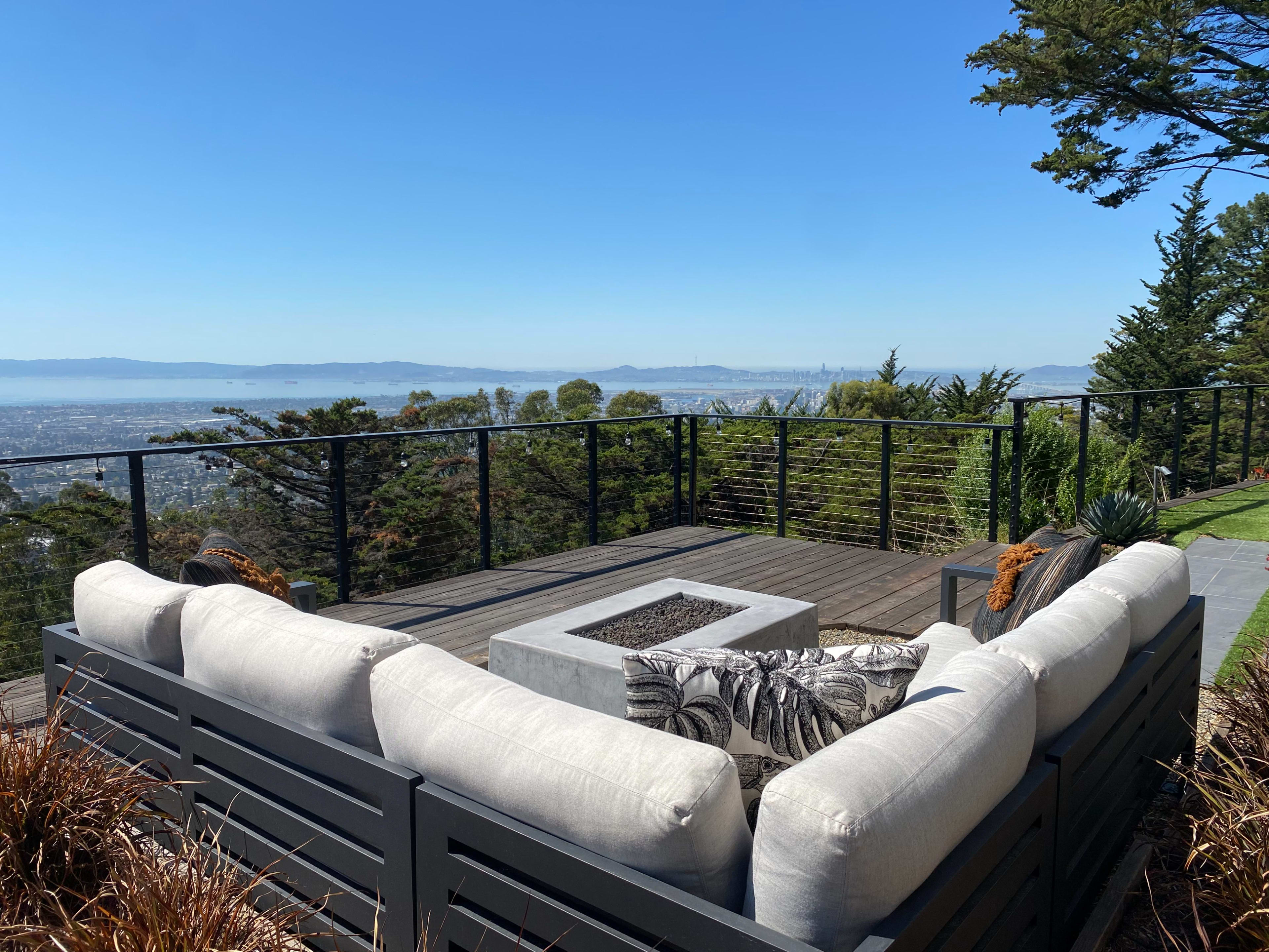 A modern outdoor lounge area with a fire pit overlooks a city and bay through a railing of trees.