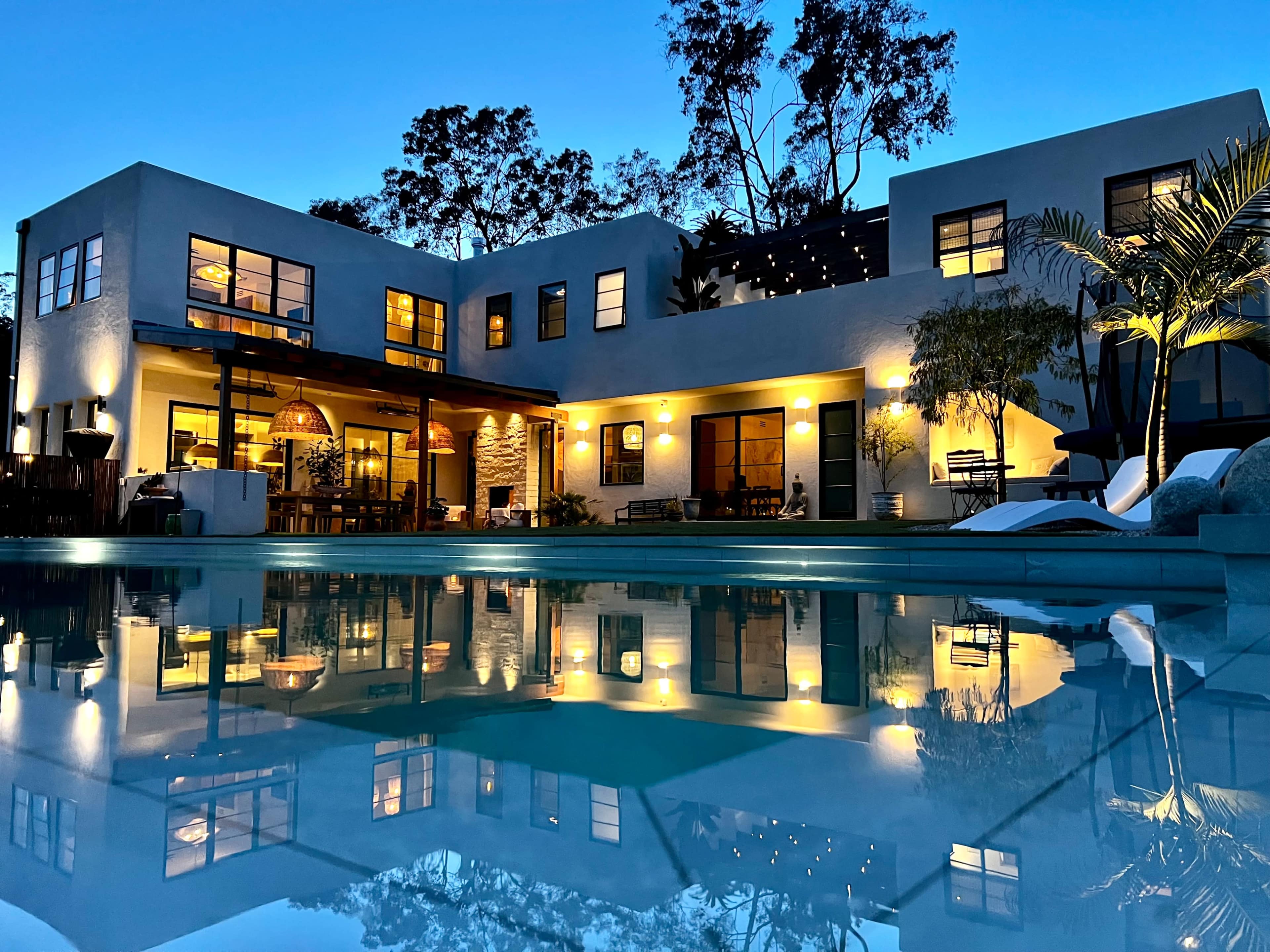 A modern two-story house with large windows reflects in the illuminated pool during twilight.