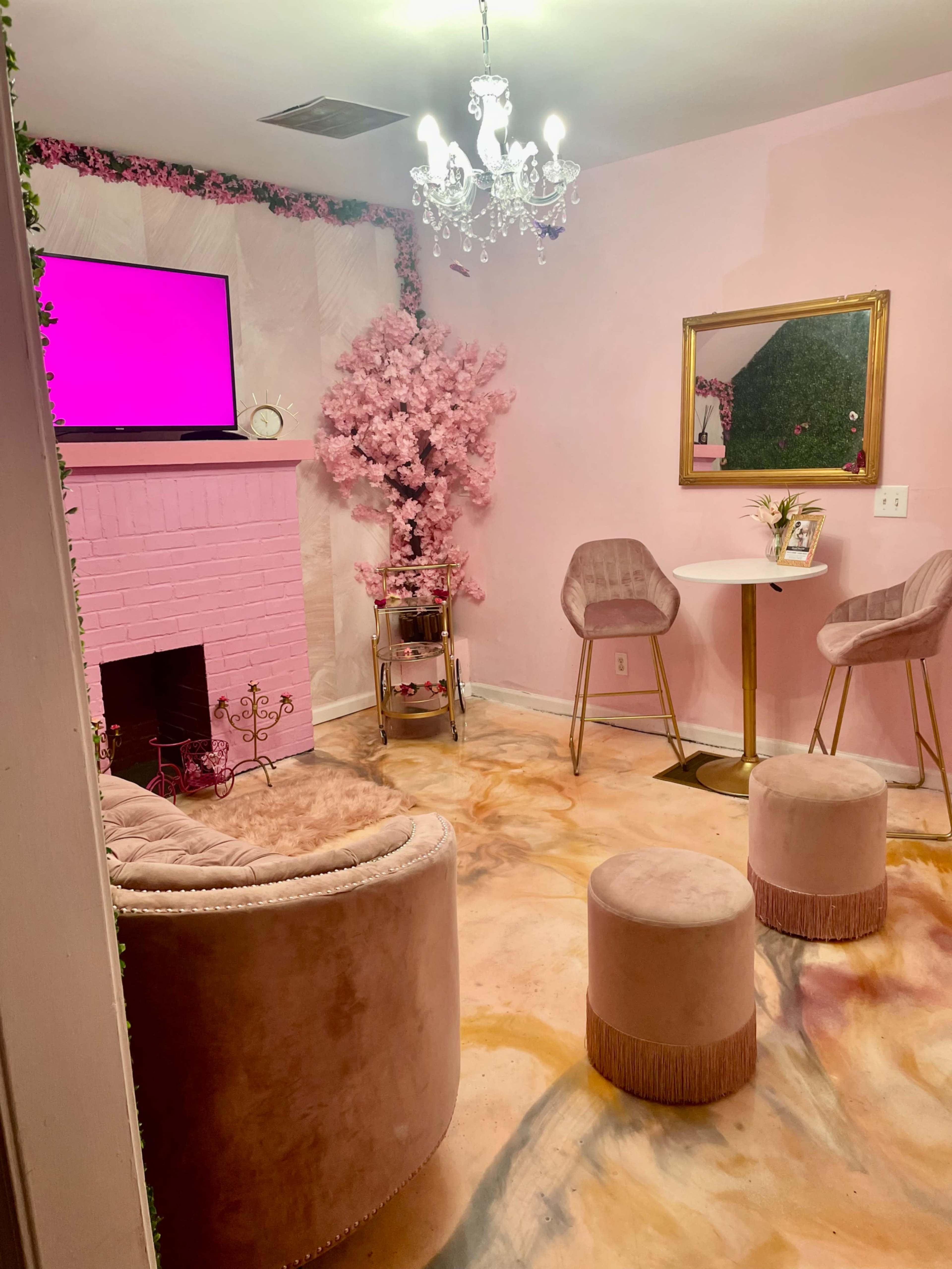 A cozy, pink-themed room featuring a TV on the wall, a chandelier, a faux cherry blossom tree, and seating arrangements of plush chairs and stools on a colorful floor.