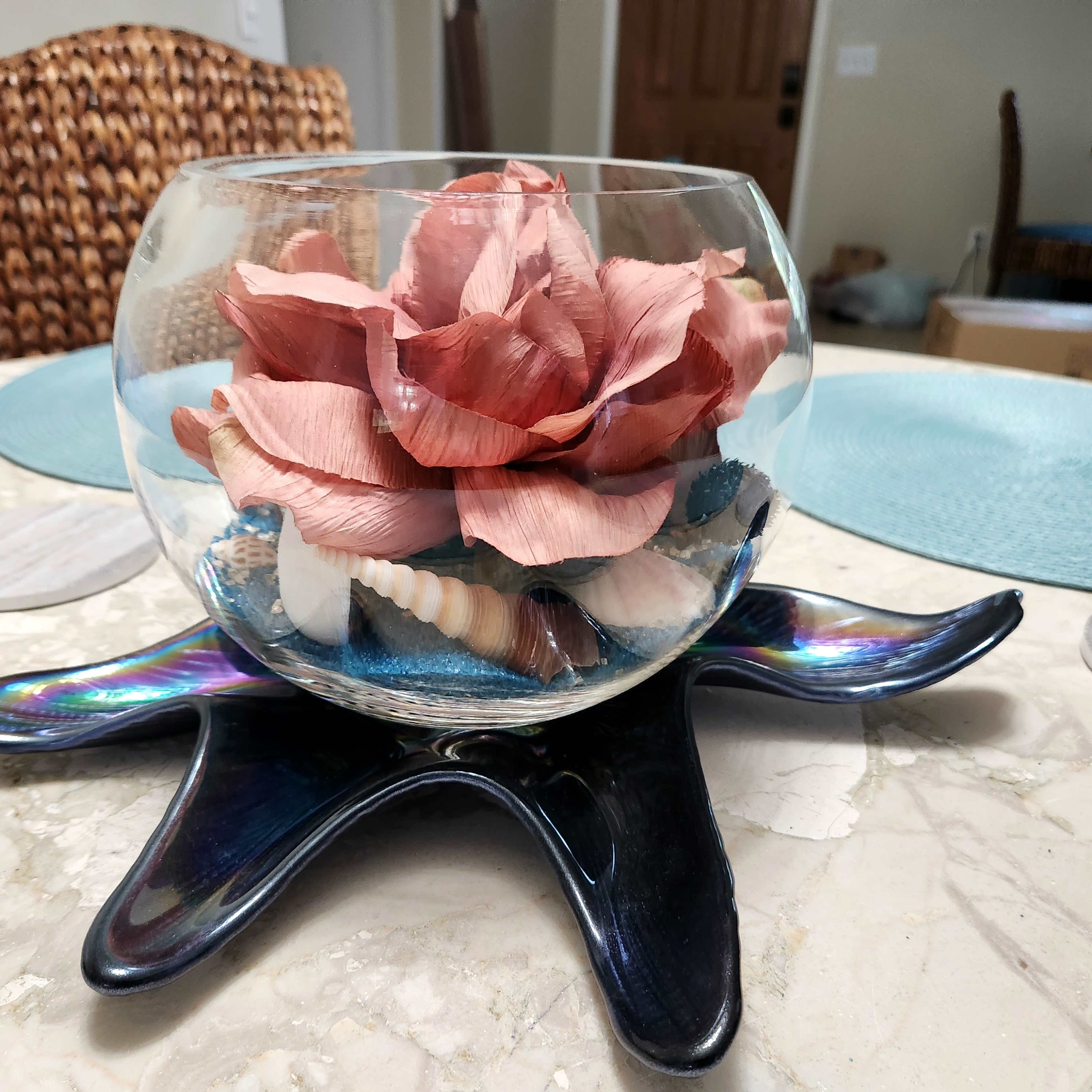 A glass bowl contains a large pink flower made of paper, surrounded by seashells and placed on a decorative shell-shaped stand on a marble table.