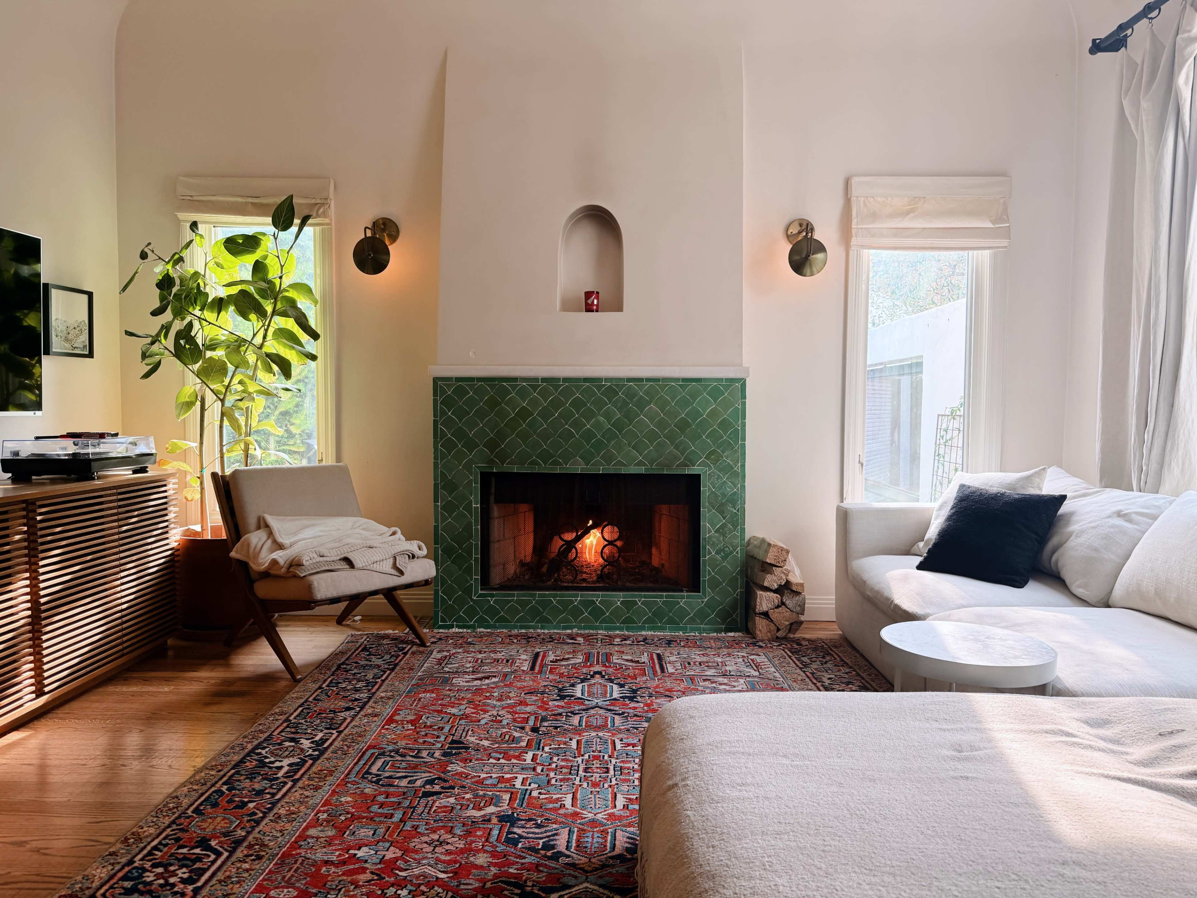A modern living room features a green tiled fireplace, a large white sectional sofa, a cozy armchair, and a patterned rug, illuminated by natural light from two windows.
