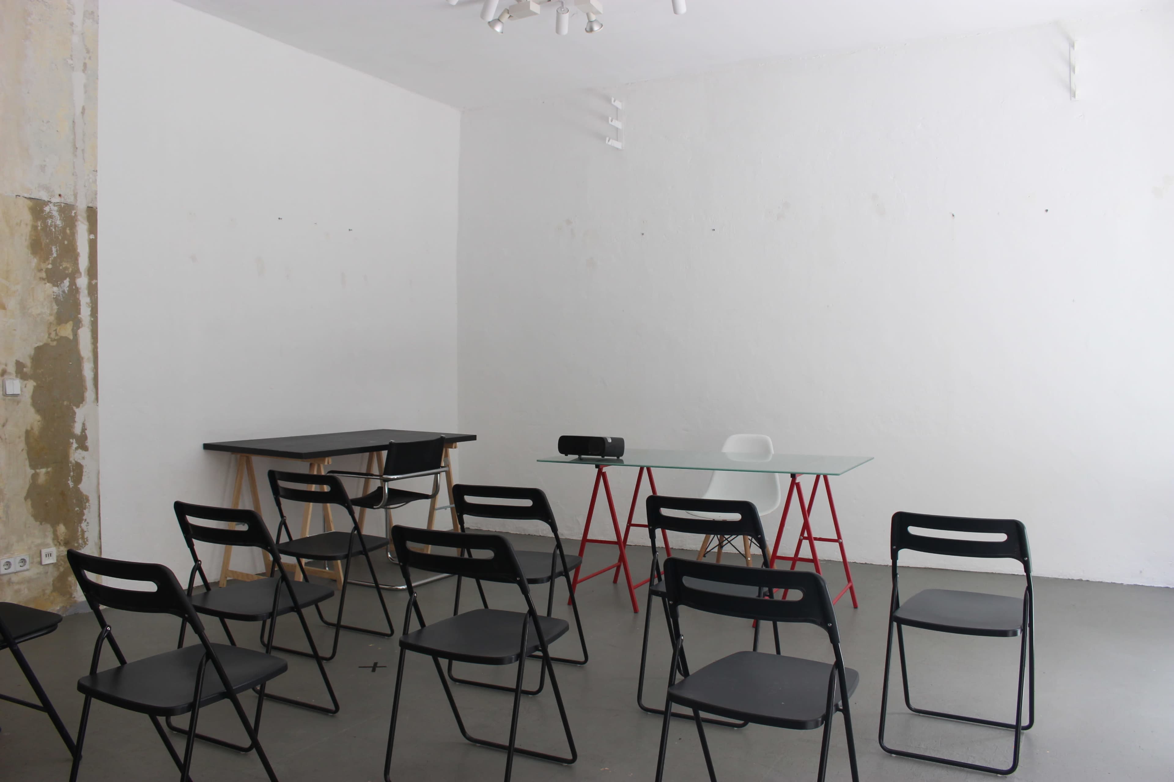 A sparsely furnished room features a glass table with red legs, a projector, and several black chairs arranged around it.