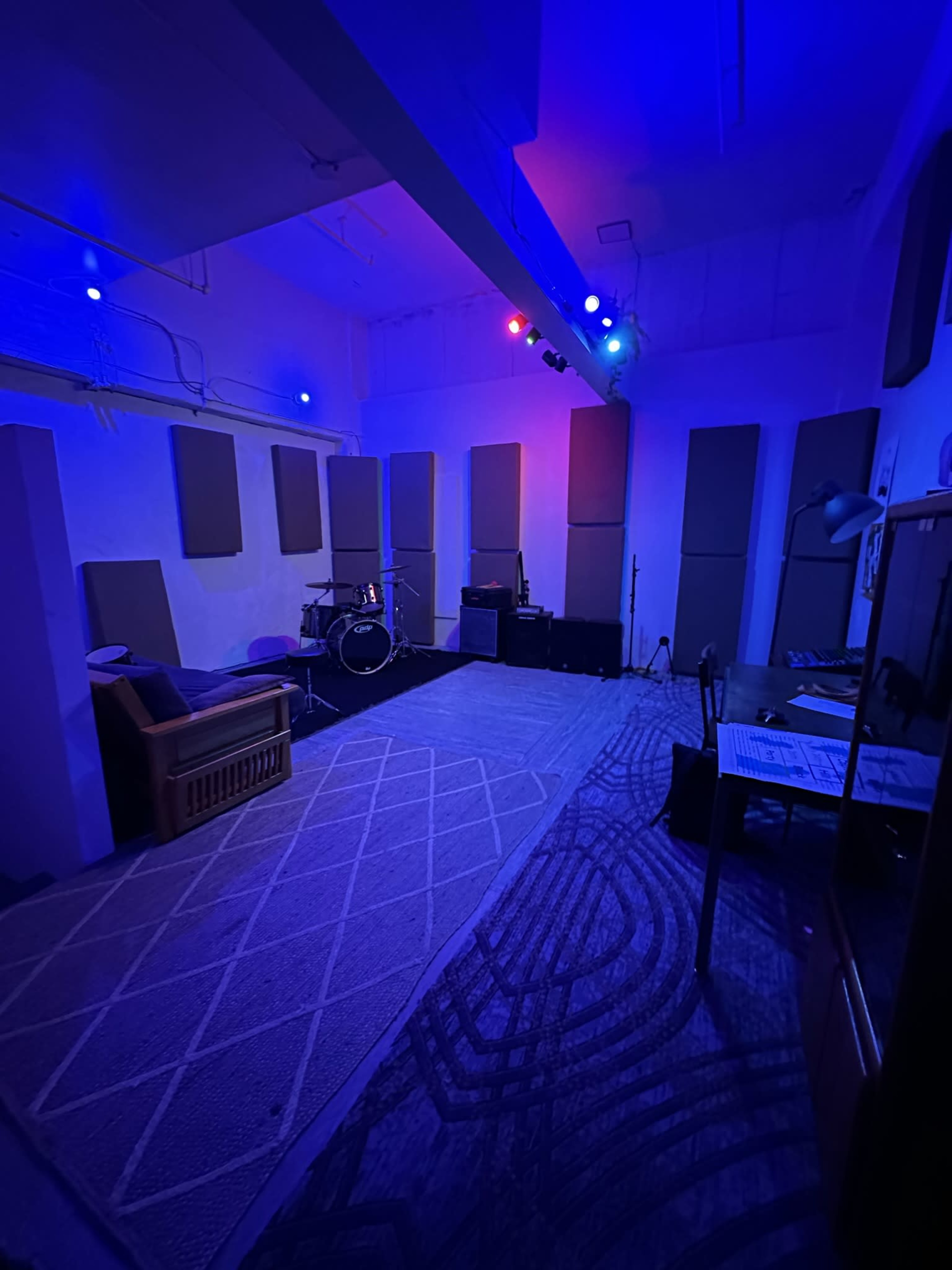 A dimly lit music studio featuring a drum set, amplifiers, soundproofing panels on the walls, and a desk with music sheets.