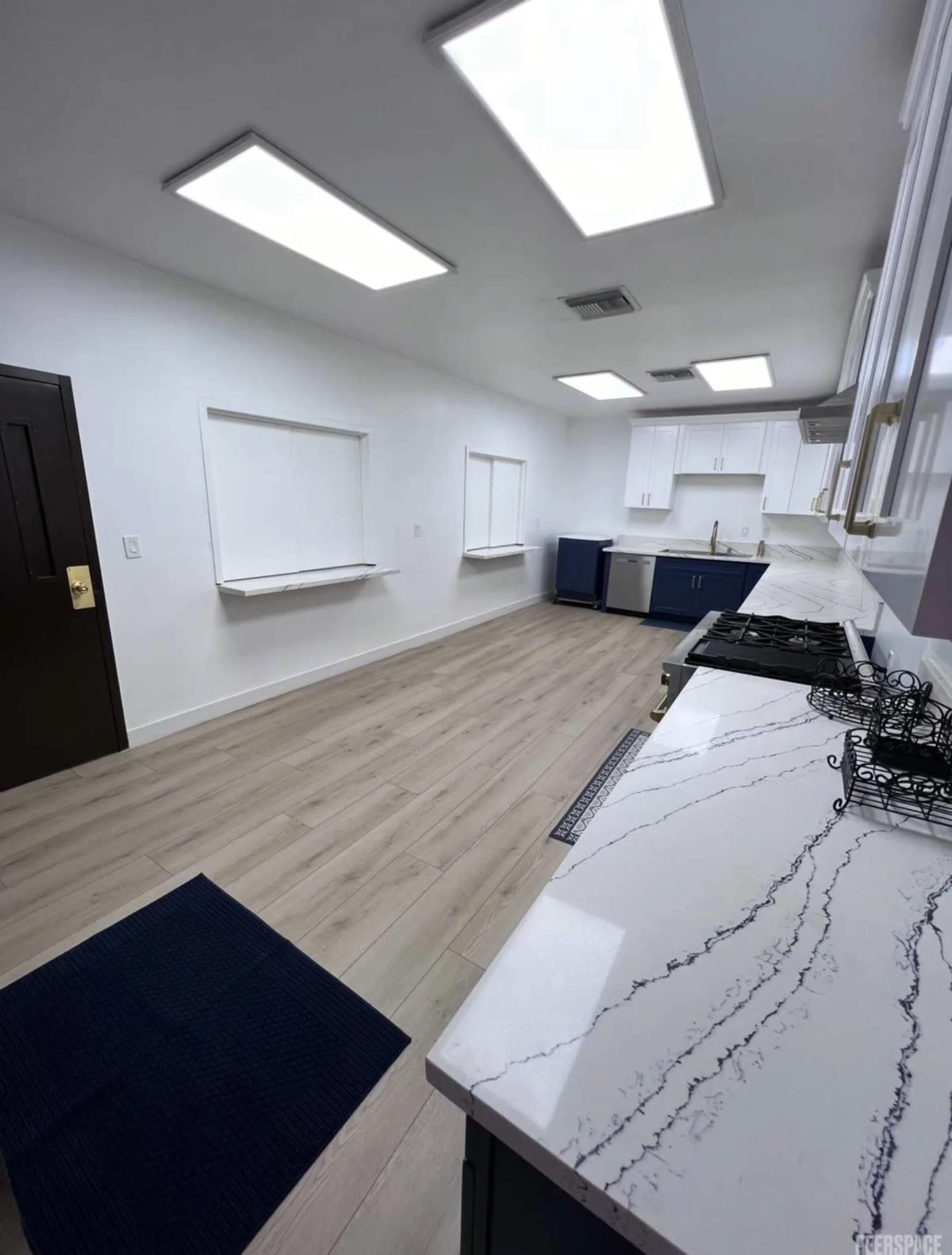 The image shows a modern kitchen with white cabinets, a marble countertop, and large windows that provide natural light.