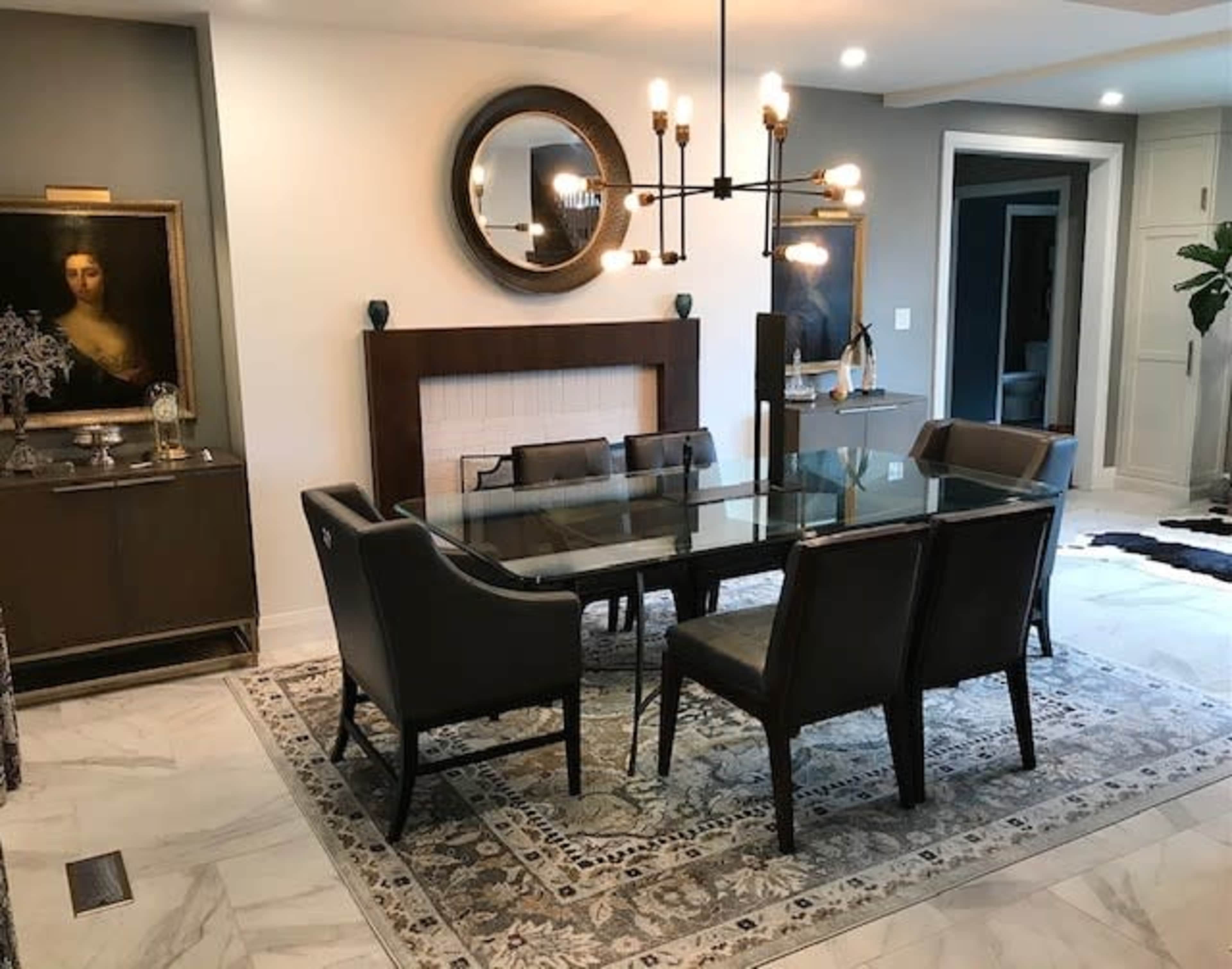 A dining room features a glass table surrounded by six upholstered chairs, with a mirror and artwork displayed on the walls.