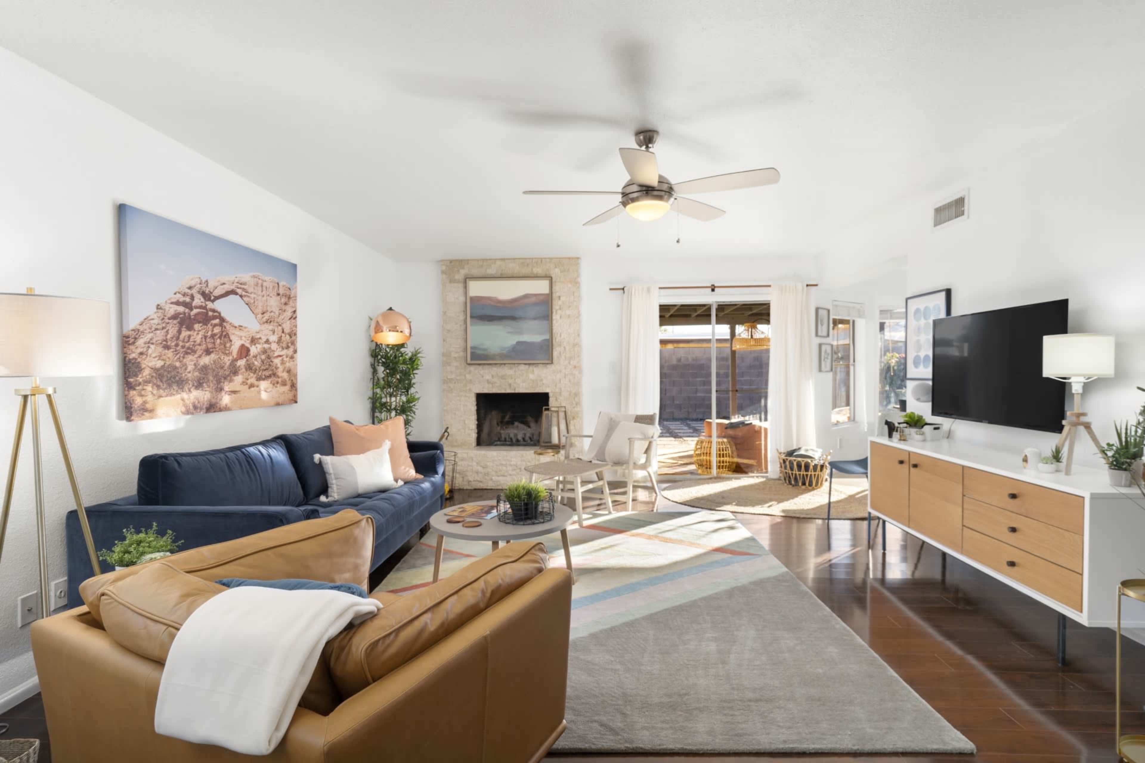 A bright living room features a blue sofa, a tan leather chair, a television unit, and a fireplace with large windows allowing natural light.