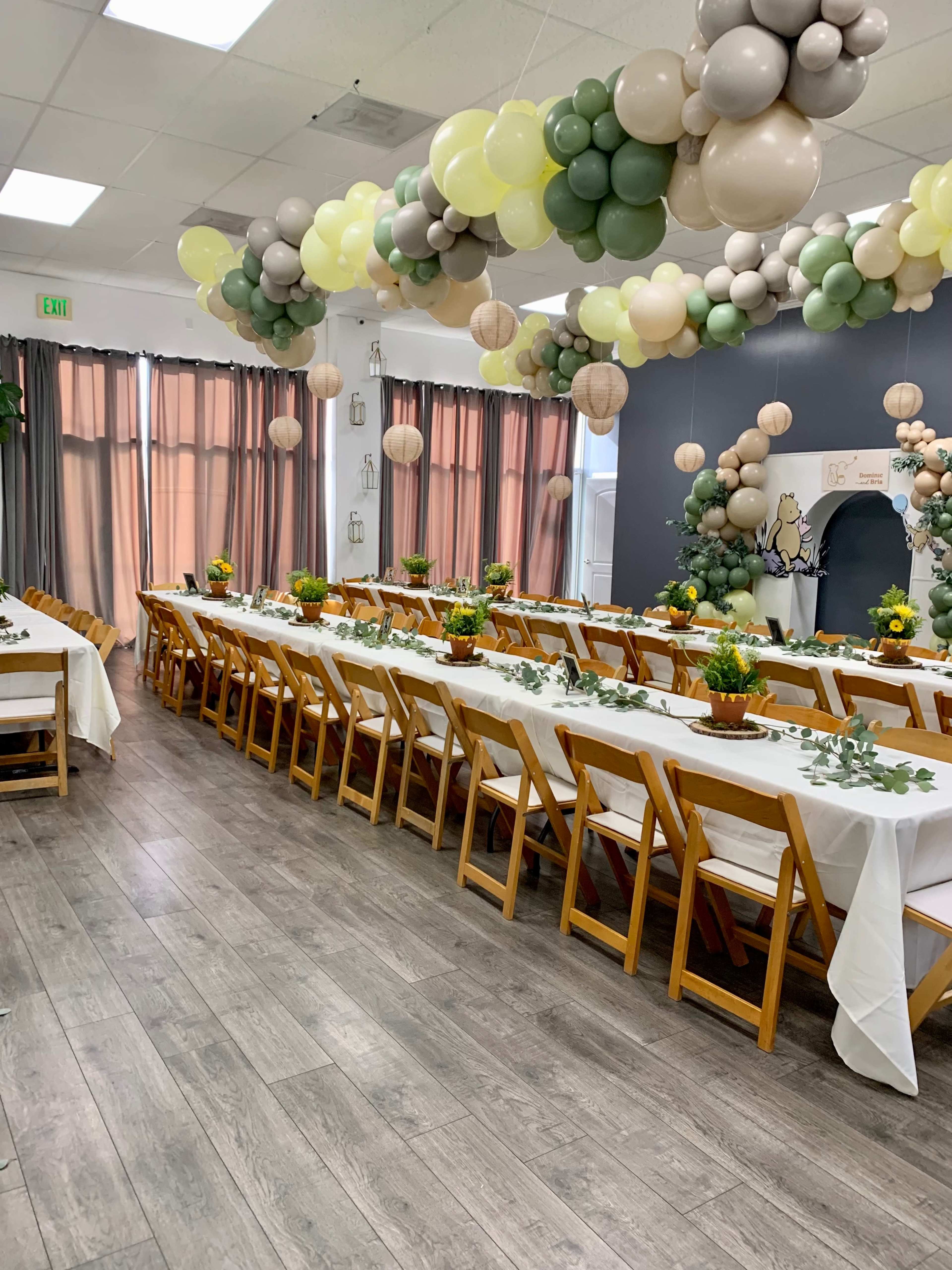 A decorated event space features long tables with white tablecloths, potted plants, and balloon garlands hanging from the ceiling.