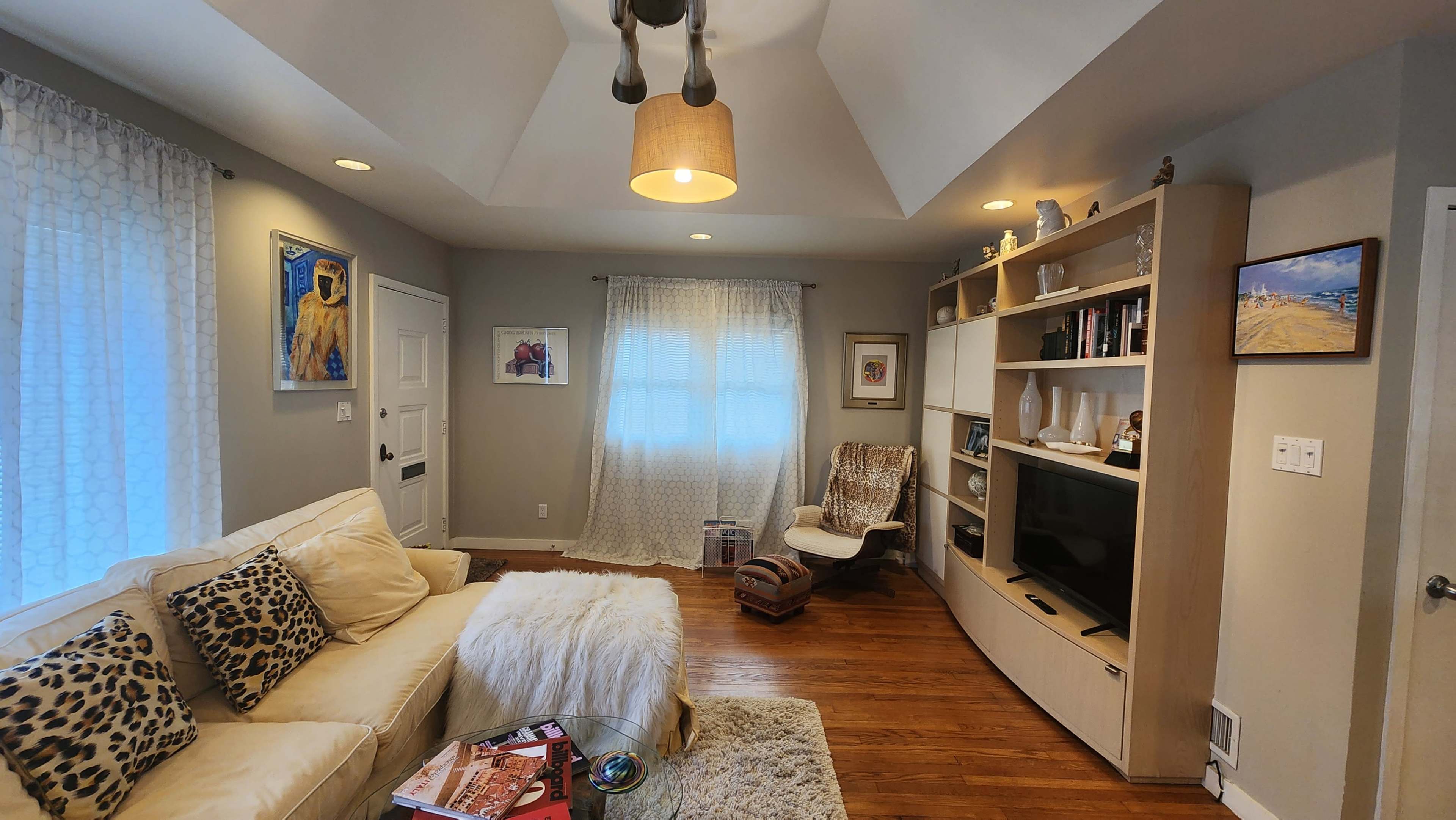 The living room features a cream-colored sofa with leopard print cushions, a wooden entertainment unit, and large windows covered with sheer curtains.