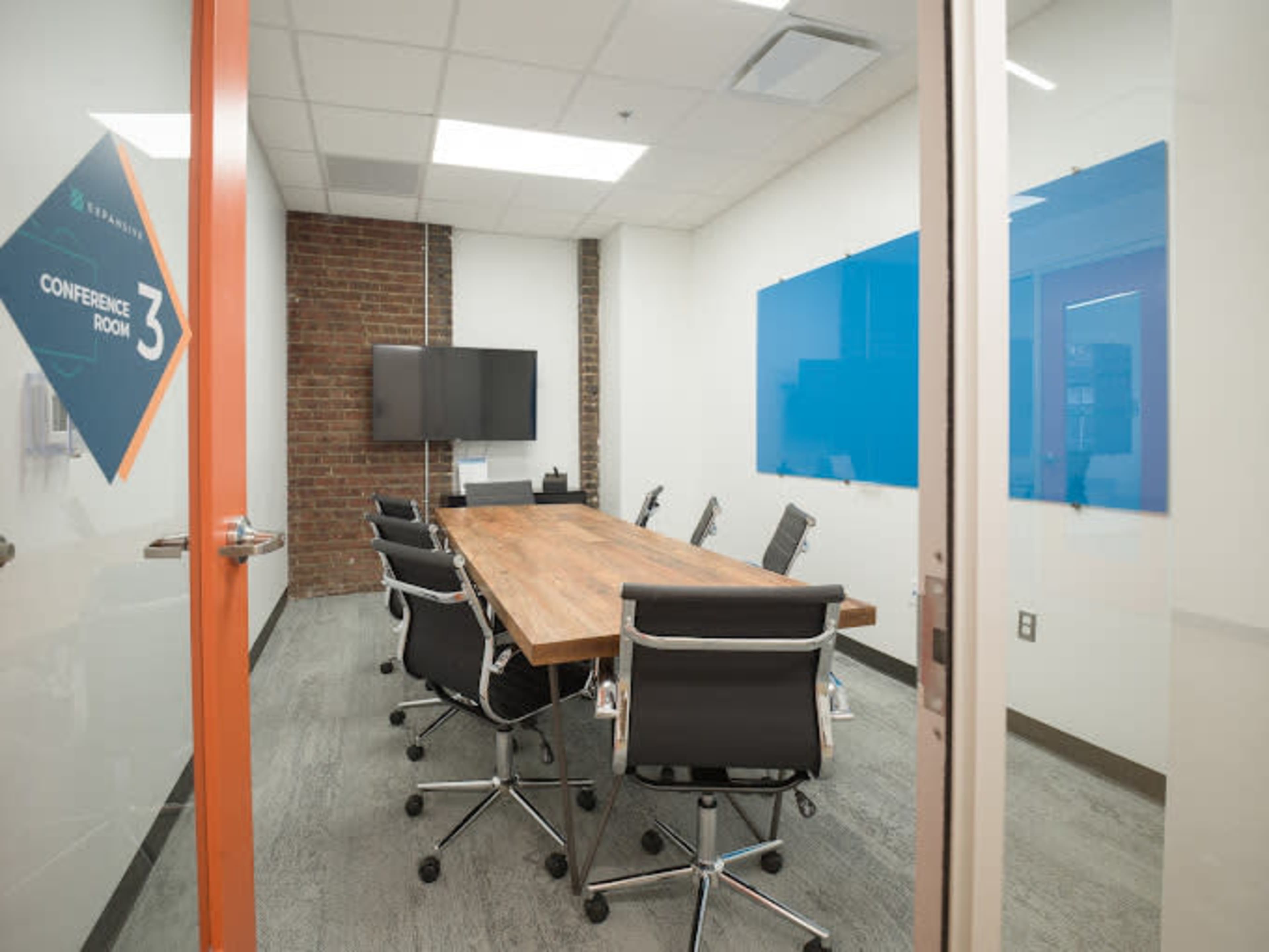 A conference room with a large wooden table, six black swivel chairs, a wall-mounted TV, and blue whiteboards on the walls.