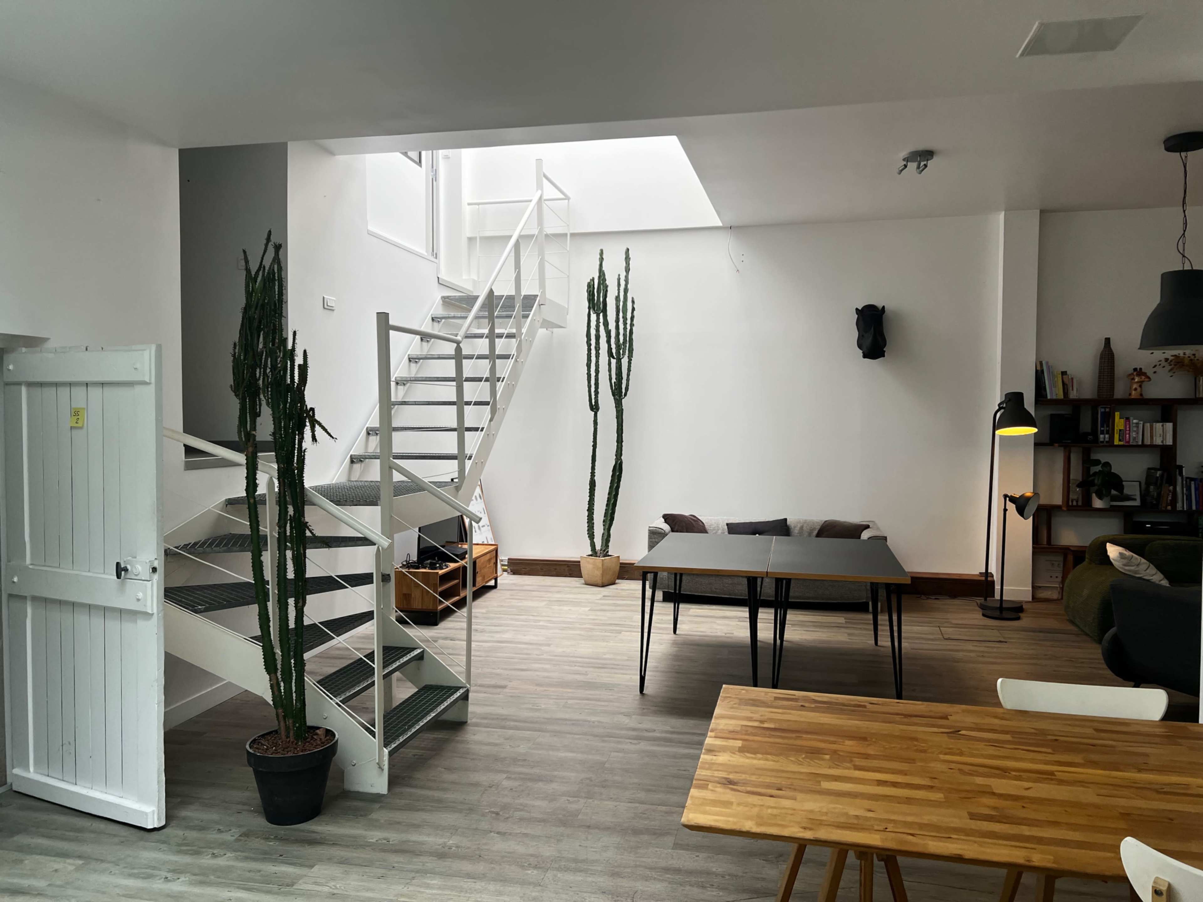 The image shows a modern, open-concept living space featuring a staircase, two tall cacti, a dining table, and a seating area with sofas.
