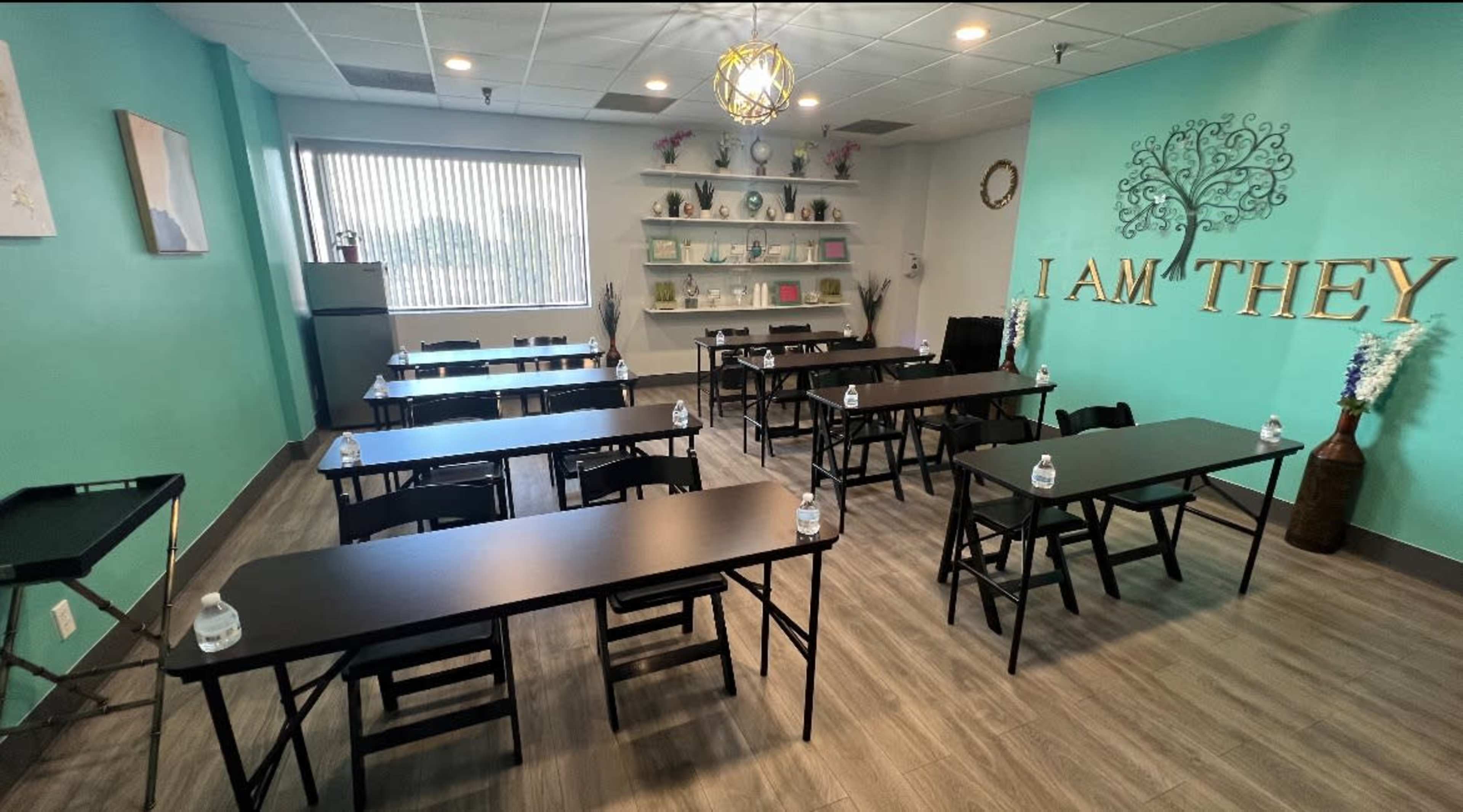 A classroom setting with several black folding tables arranged in rows, each with a bottle of water, and a wall displaying the text "I AM THEY" alongside shelves filled with various items.