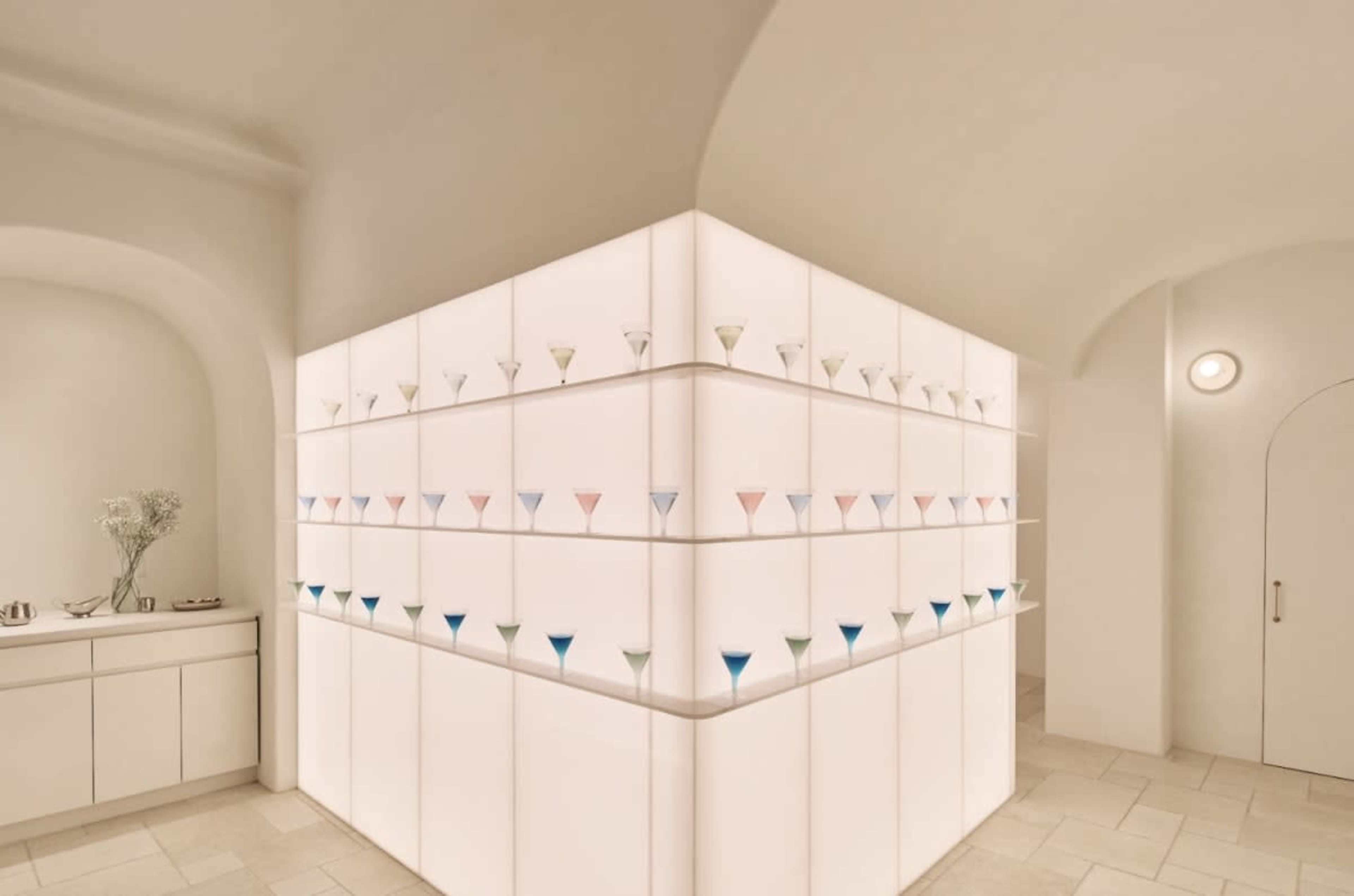 The image shows a modern, minimalist interior with a corner display featuring colorful glass cups arranged on illuminated shelves.