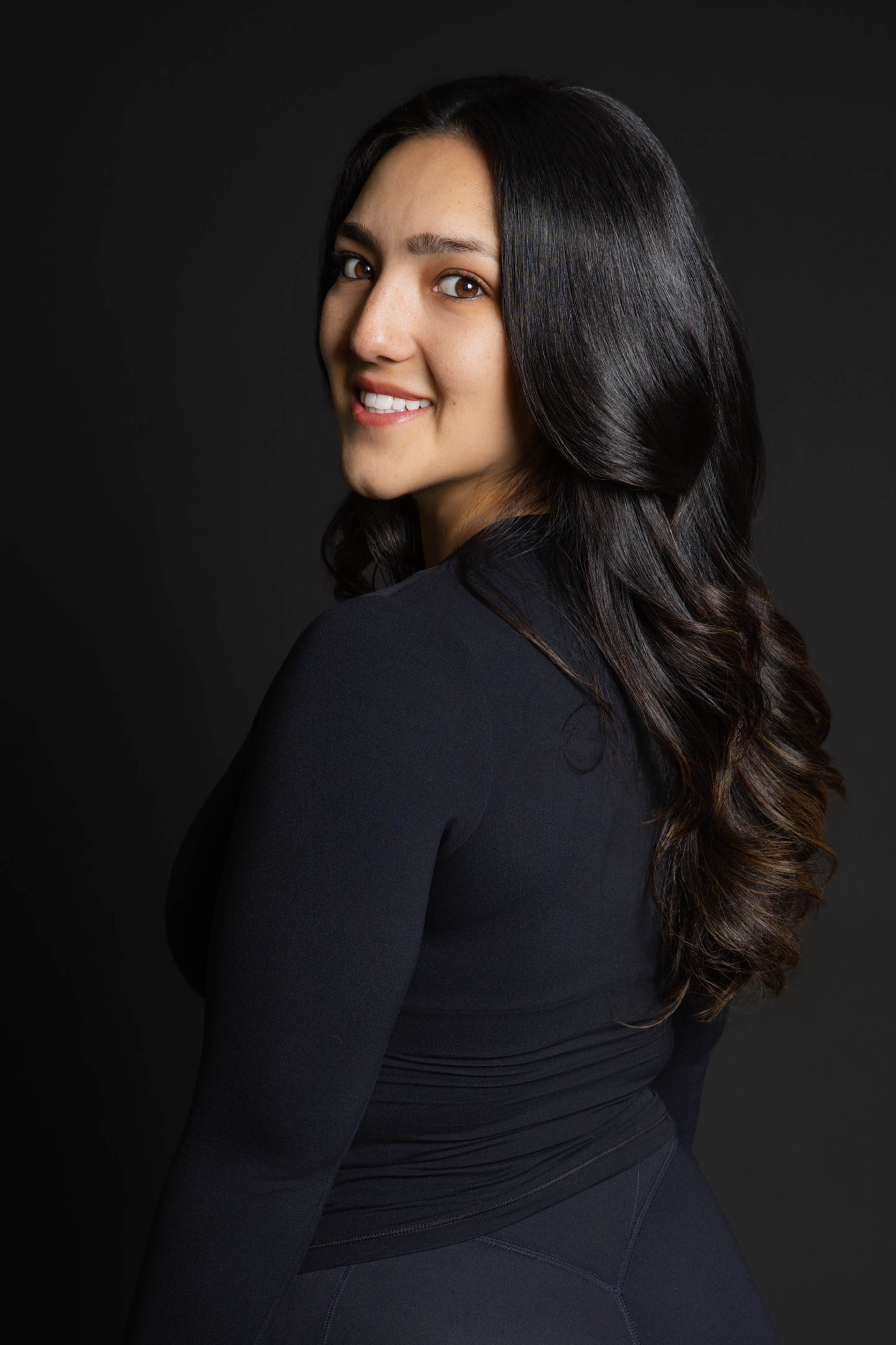 A woman with long, dark hair smiles while posing against a dark background.