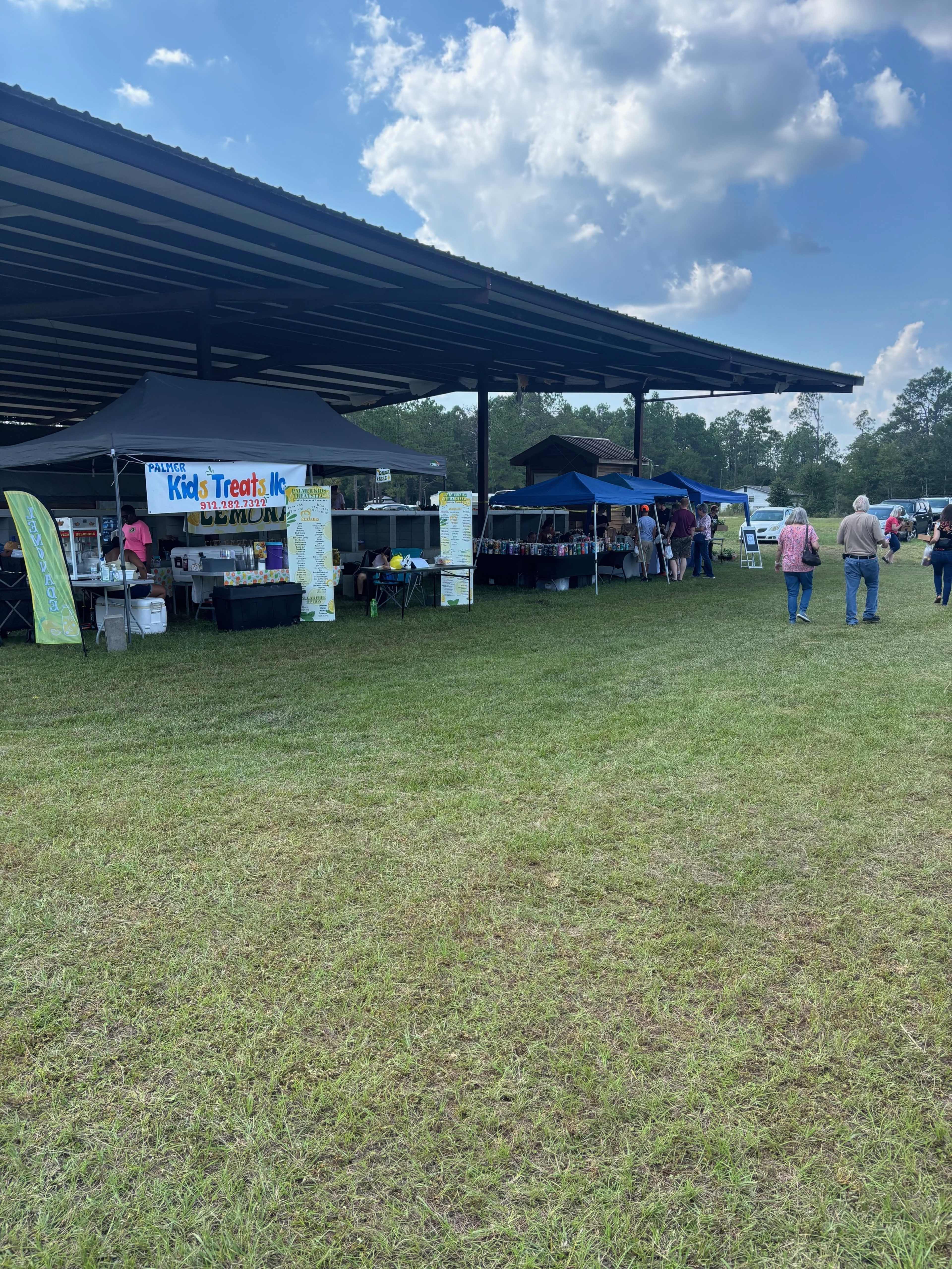 The image shows a grassy area with several vendor tents set up under a covered structure during a fair or outdoor event.
