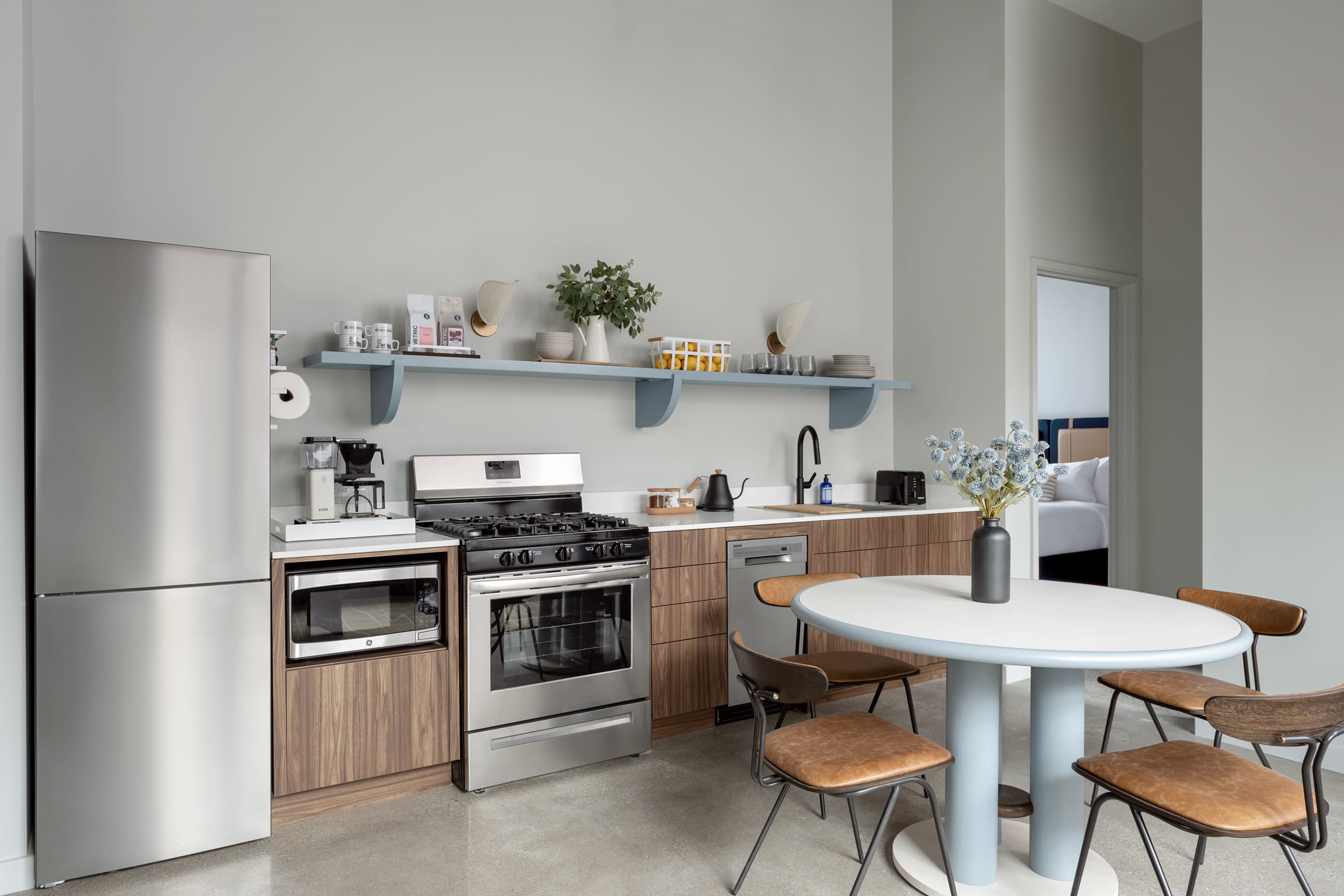 A modern kitchen features stainless steel appliances, wooden cabinetry, a circular dining table, and a shelf holding various kitchen items against a pale wall.