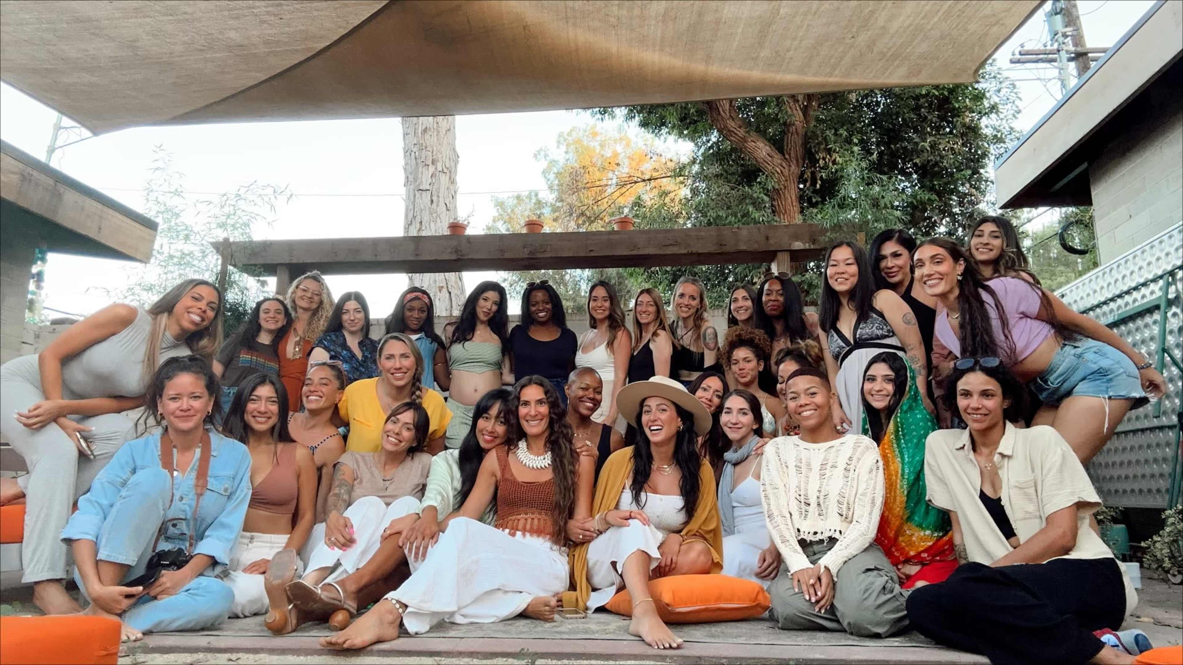 A large group of women is gathered together outdoors on a patio, sitting and standing in front of a shaded area with greenery in the background.