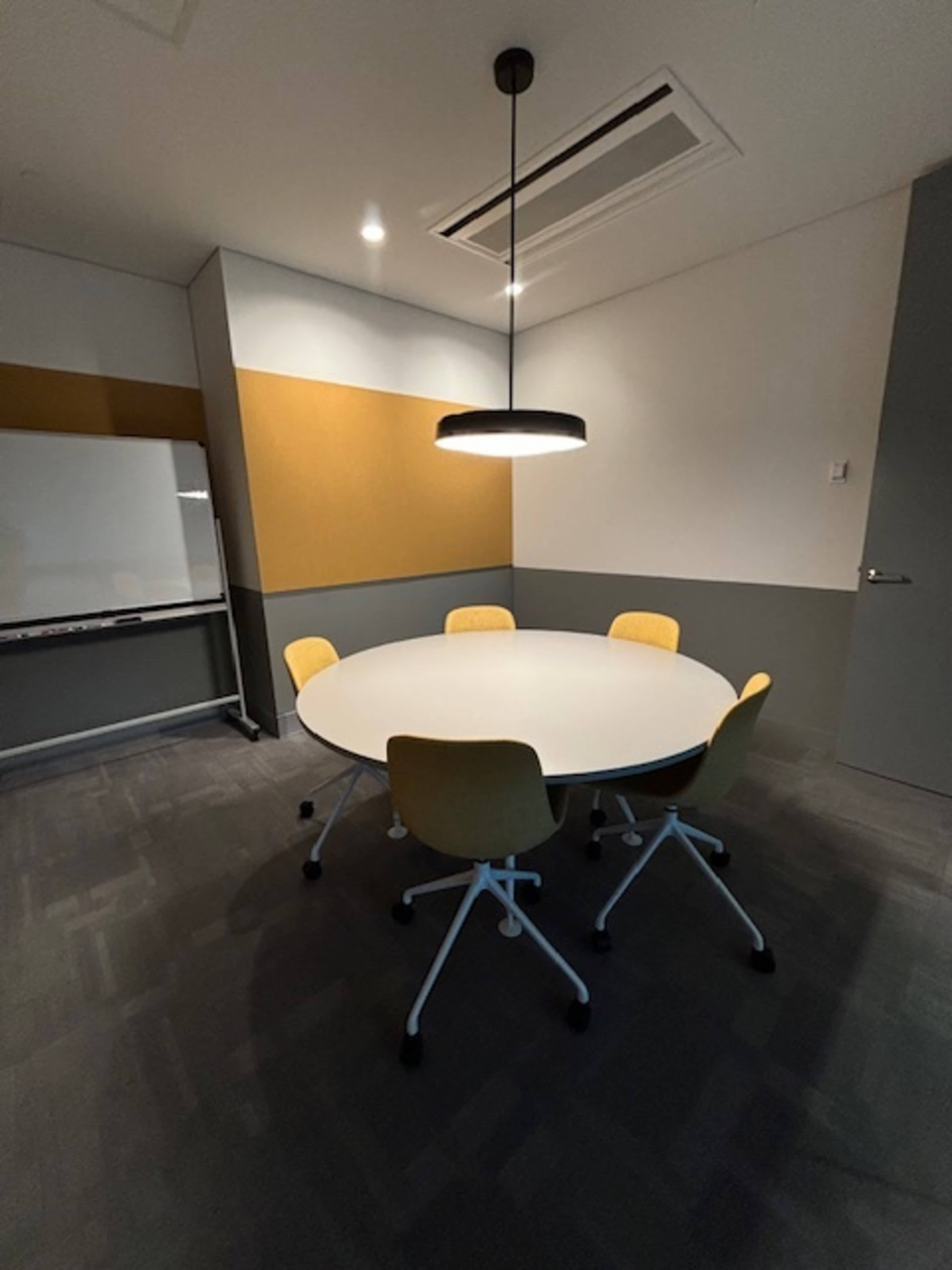 A round table with four yellow chairs is situated in a meeting room with gray walls and a whiteboard.