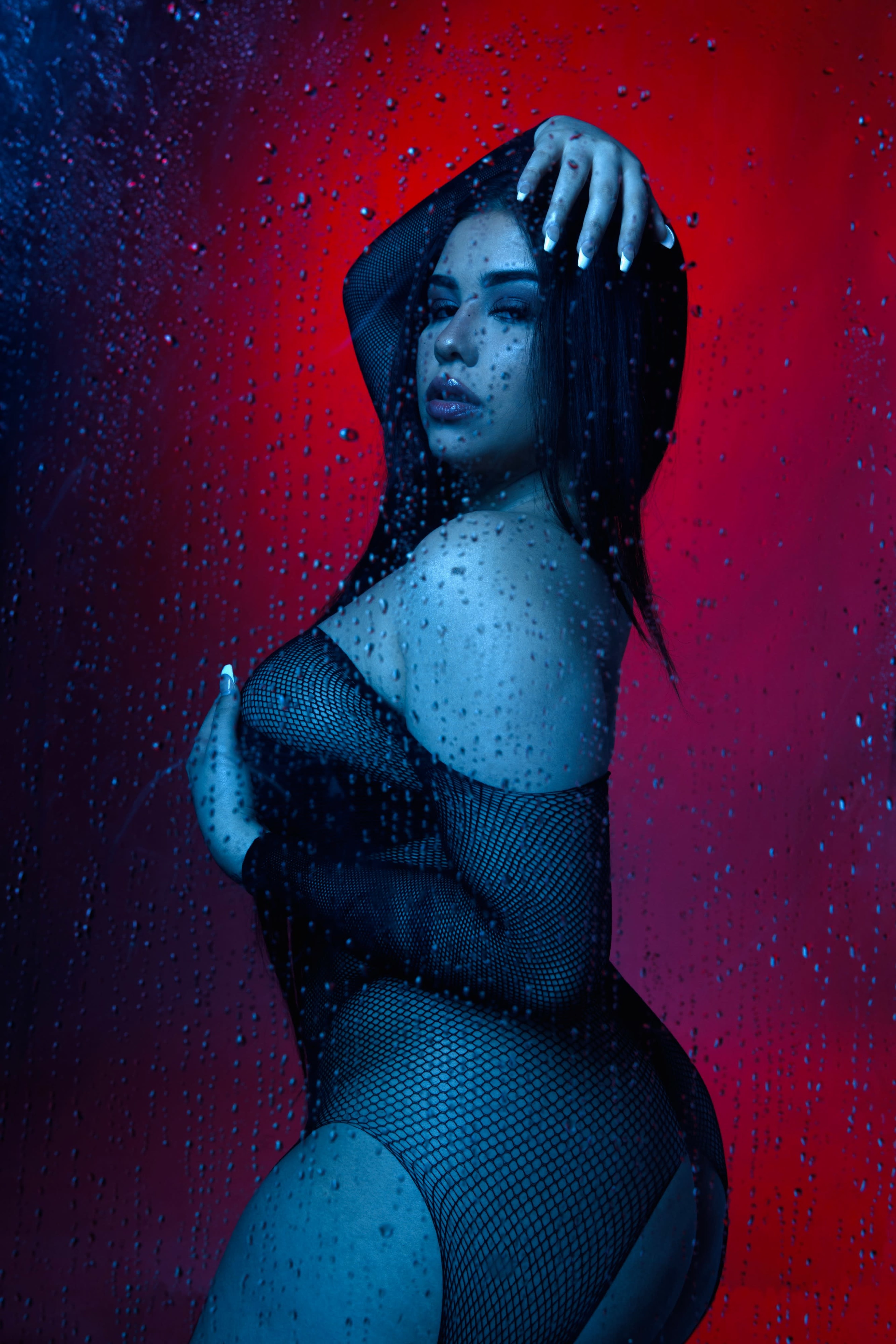 A person poses behind a glass surface covered in water droplets, with a contrasting red and blue background.