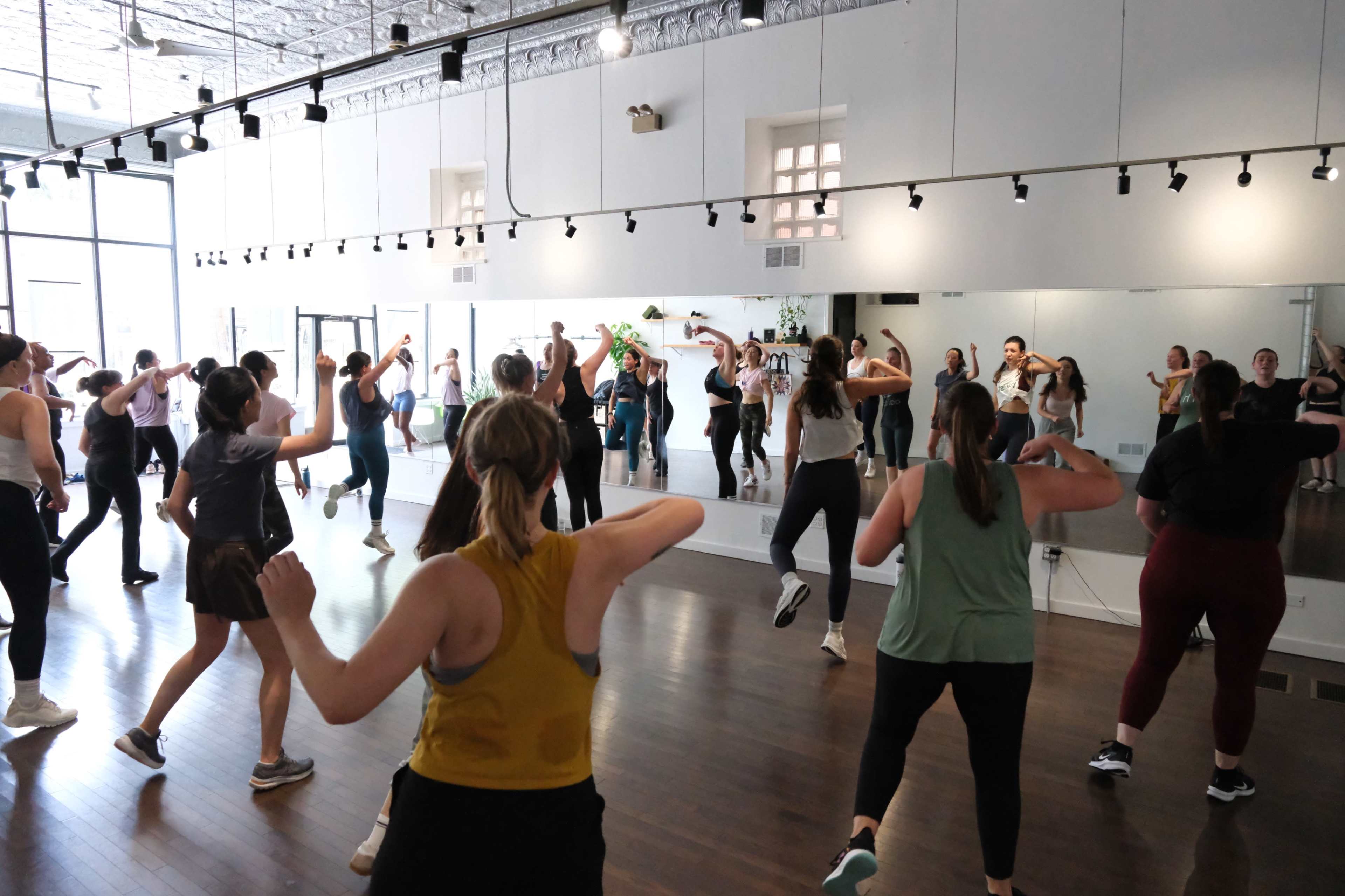 A group of people is participating in a dance class in a studio with mirrored walls and wooden flooring.