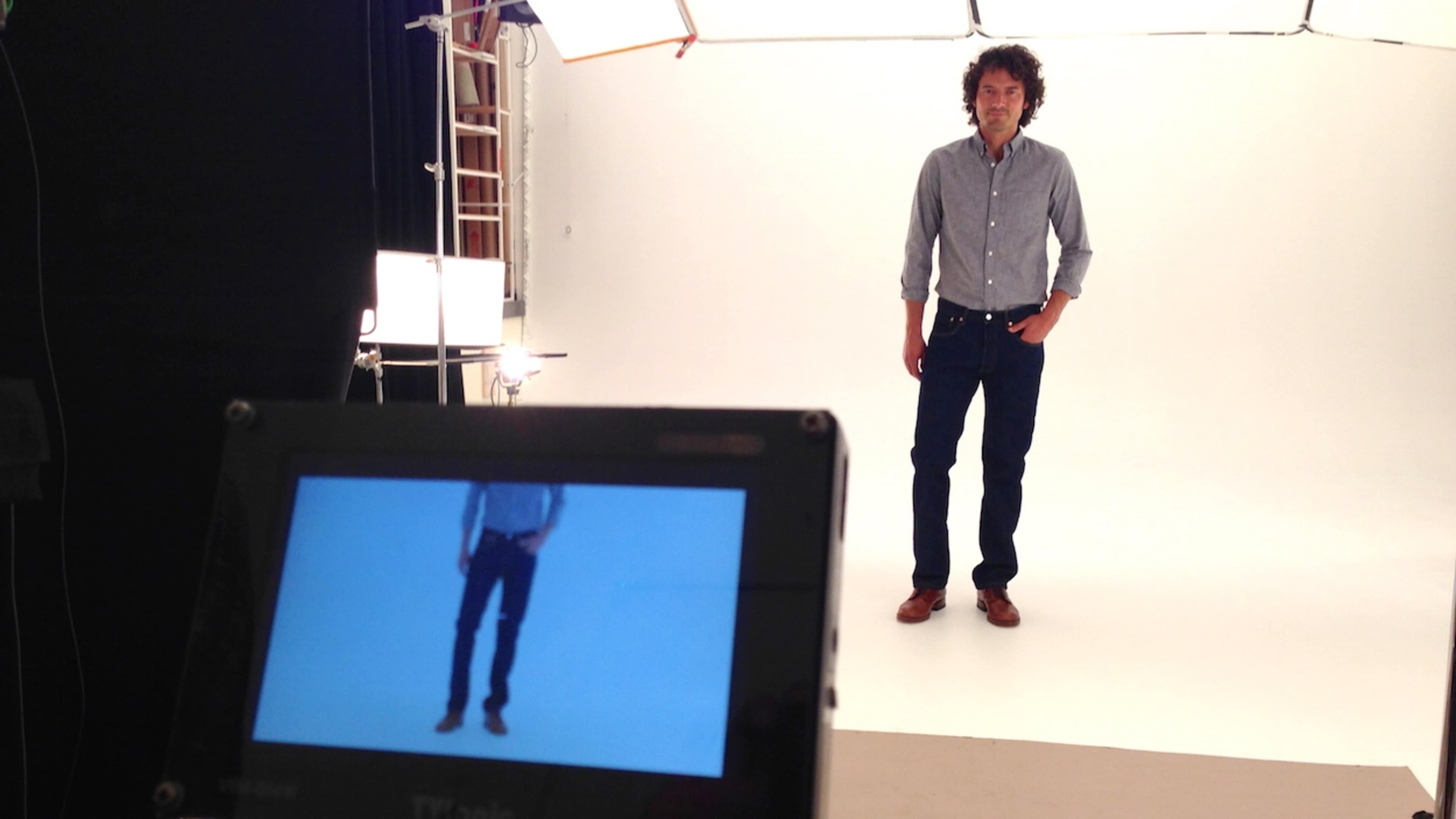 A man stands posing in a studio while being photographed, with a monitor displaying his image in front of him.
