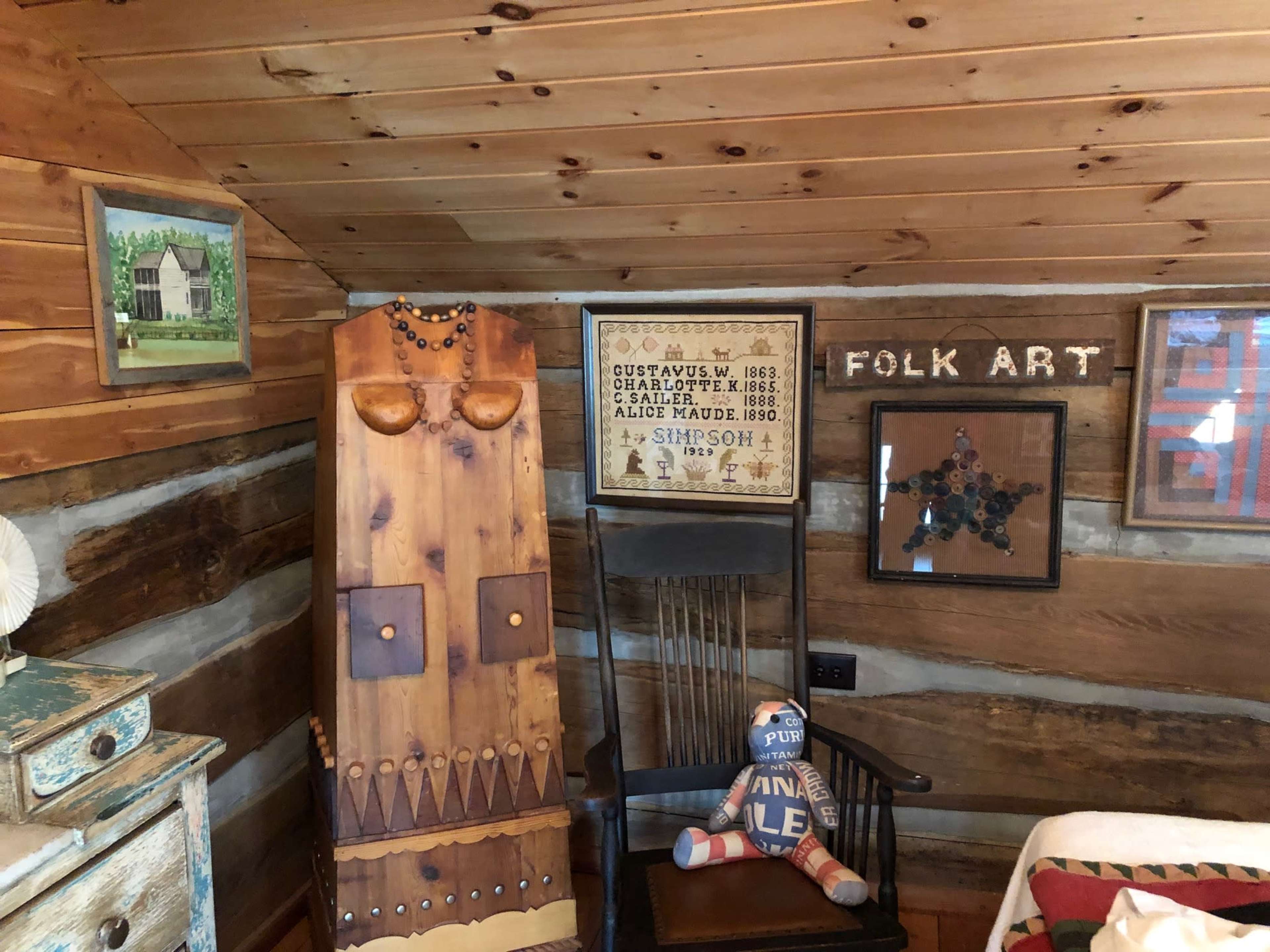 A rustic log cabin interior featuring a wooden chair, a folk art display on the wall, and a wooden sculpture resembling a figure.