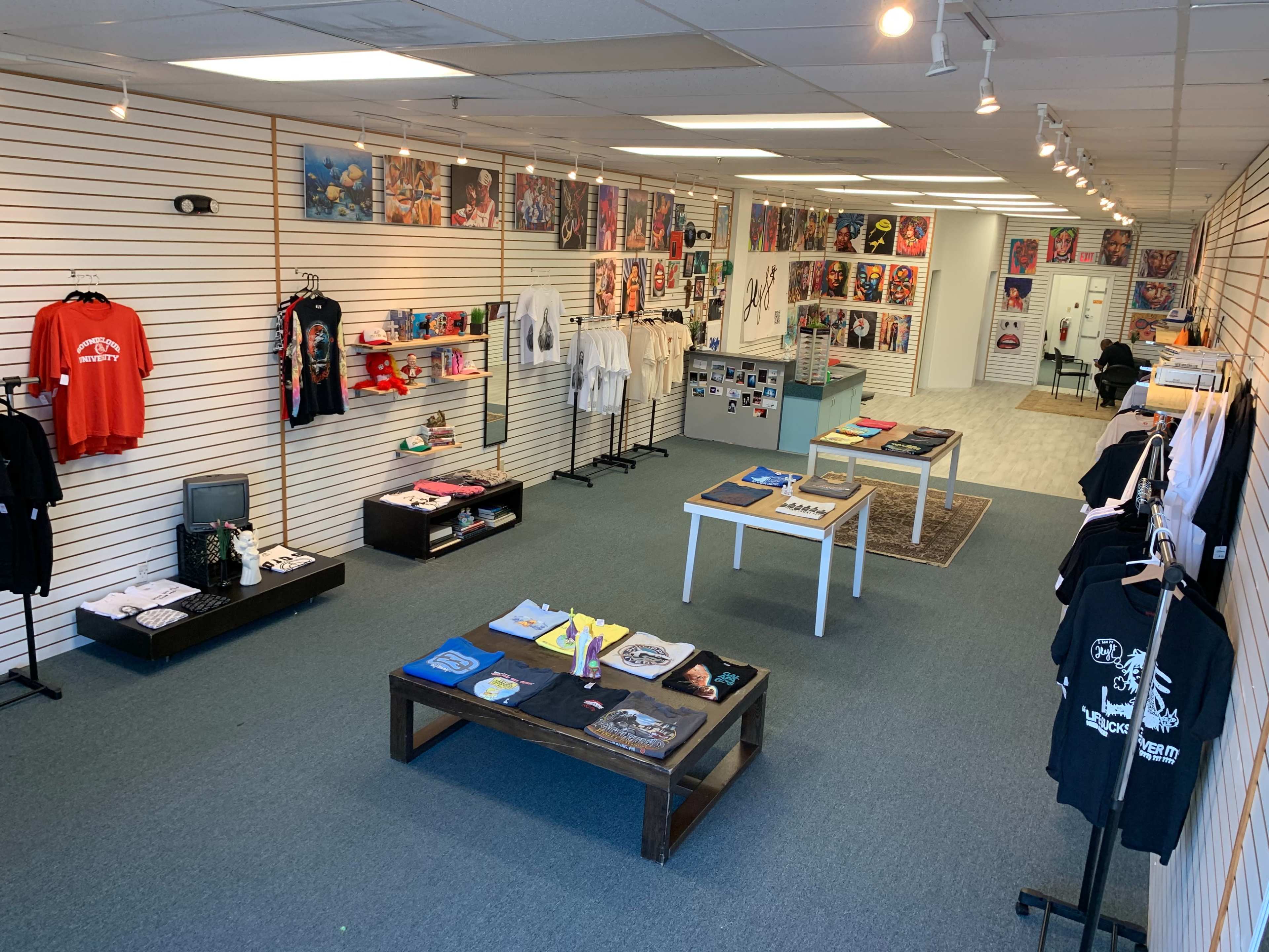 The image shows an apparel store featuring clothing displays on racks, a central table with various folded shirts, and walls adorned with colorful artwork and posters.