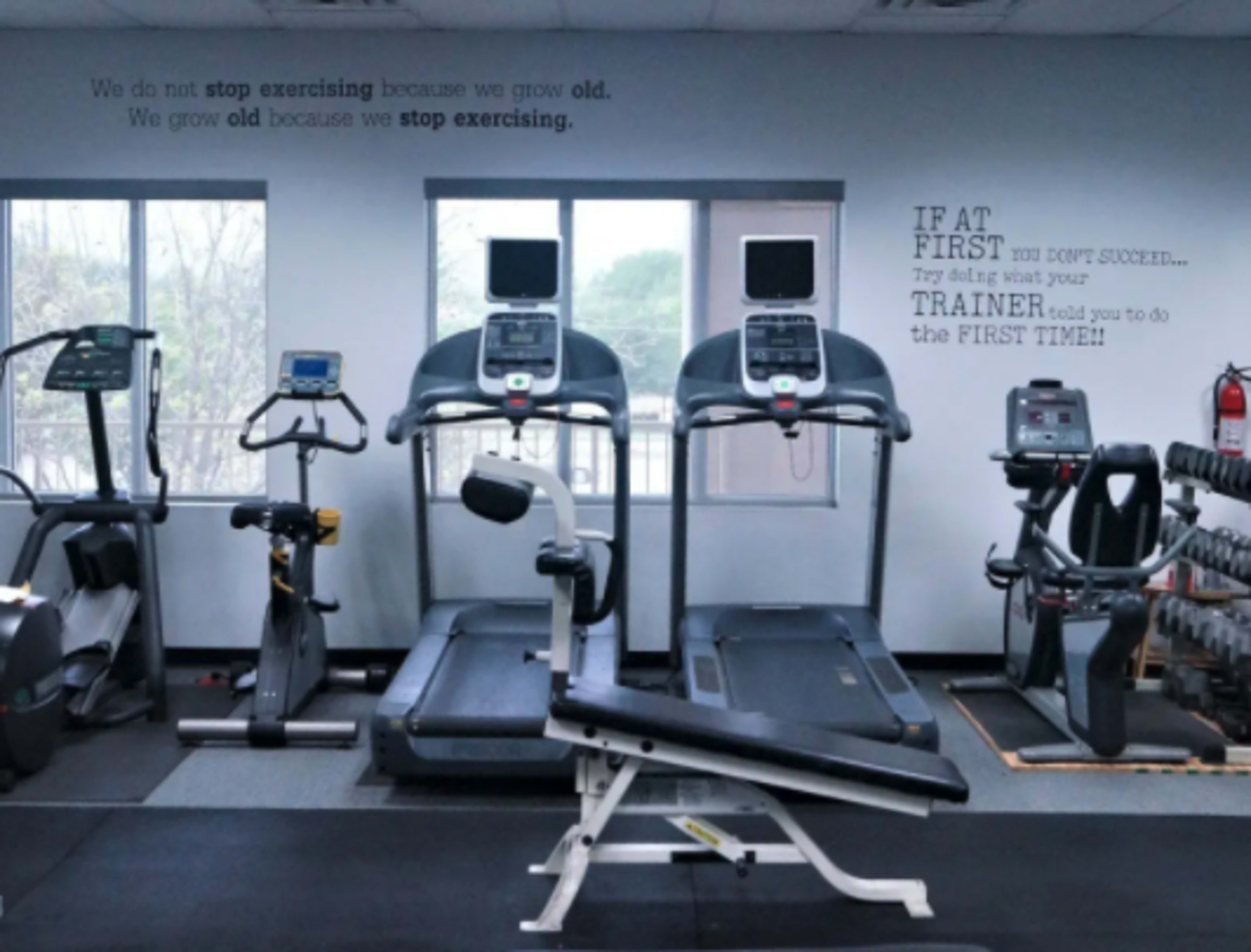 A fitness area with several exercise machines, including treadmills and a weight bench, against a wall with motivational quotes.