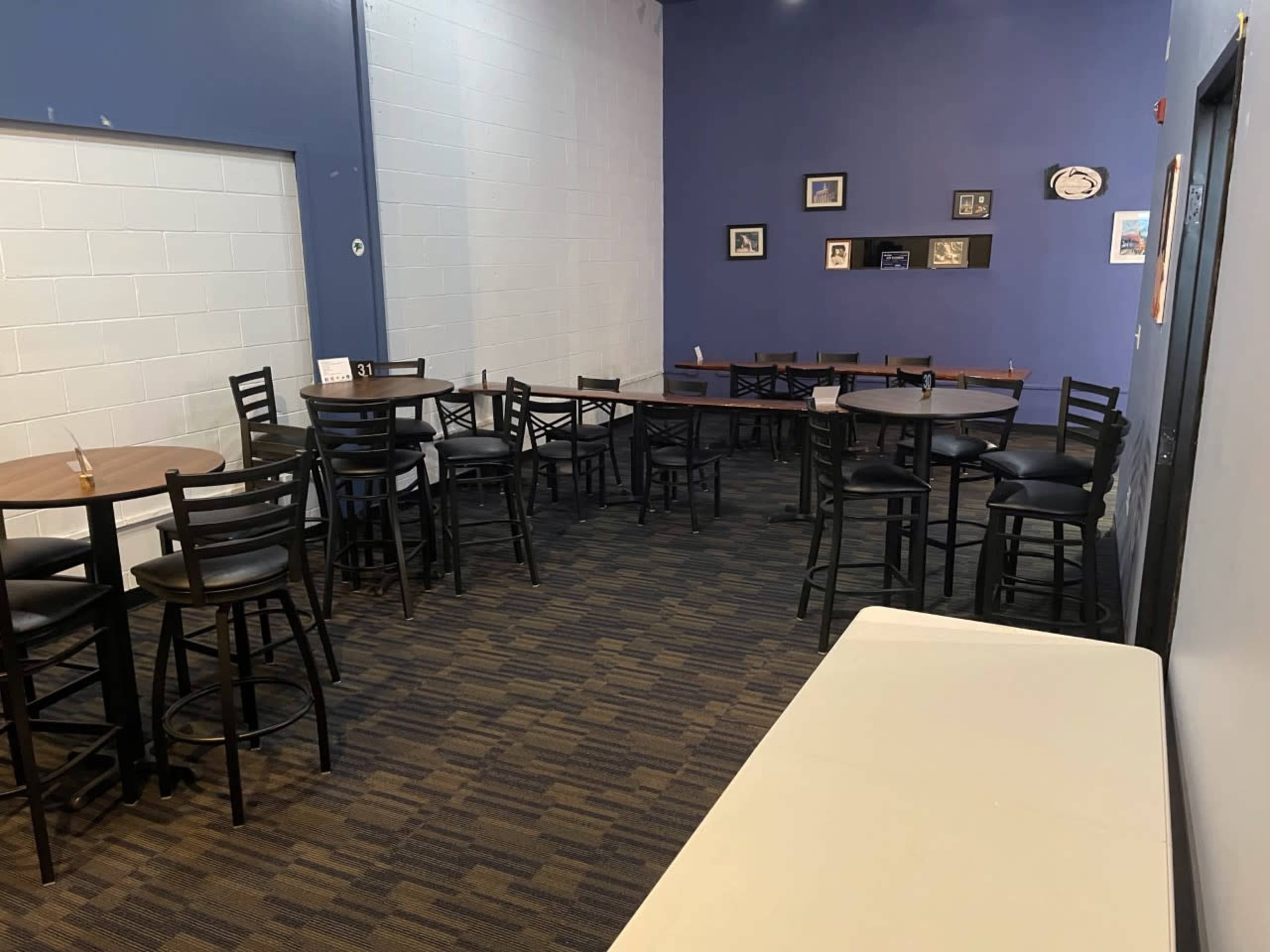 A small, partially empty room features several tall tables and chairs arranged on a carpeted floor, with blue walls and a few framed pictures on display.