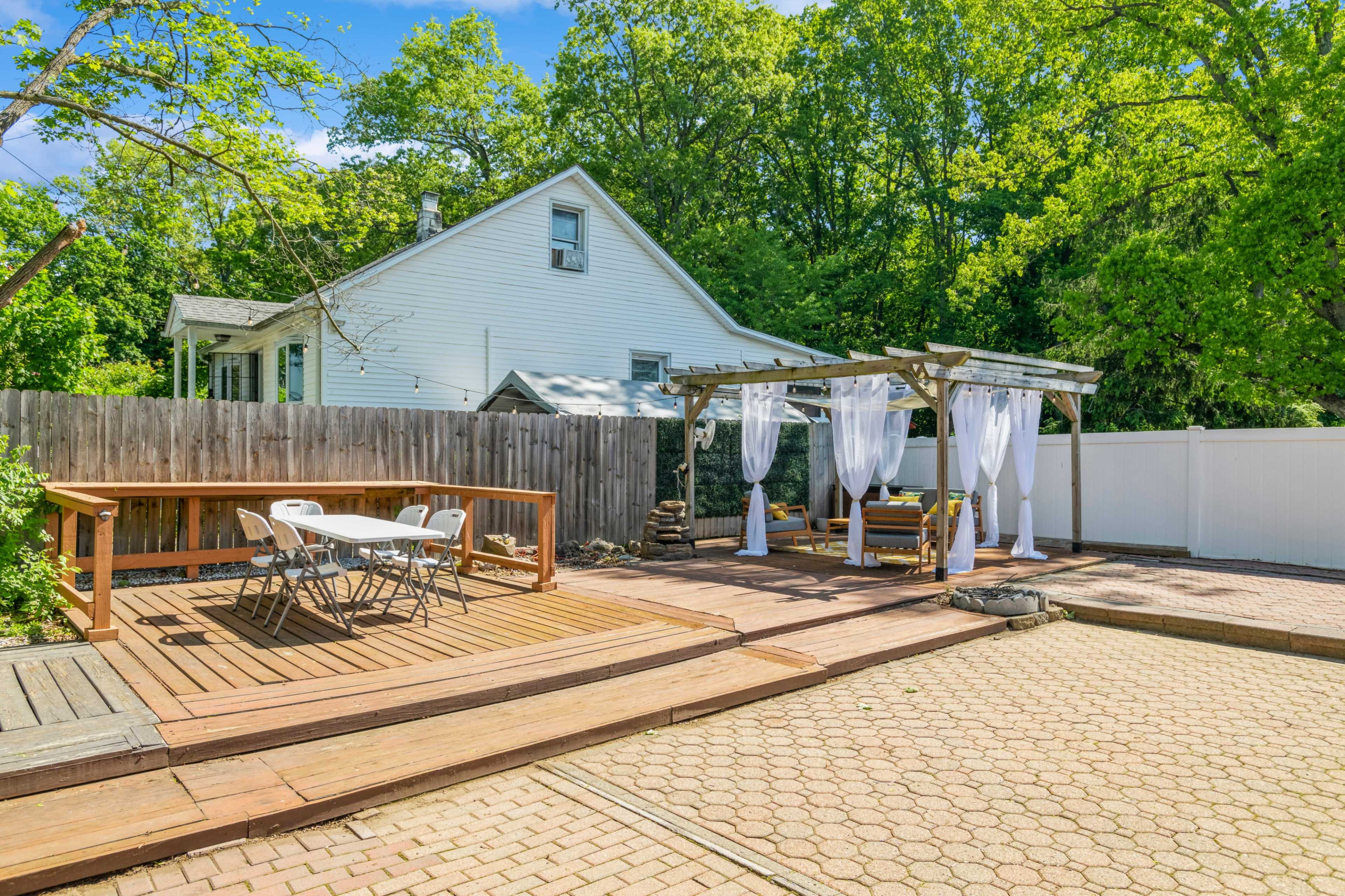 The image shows a backyard with a wooden deck, a pergola with white drapes, a seating area, and a table set for dining, surrounded by greenery and a fenced perimeter.