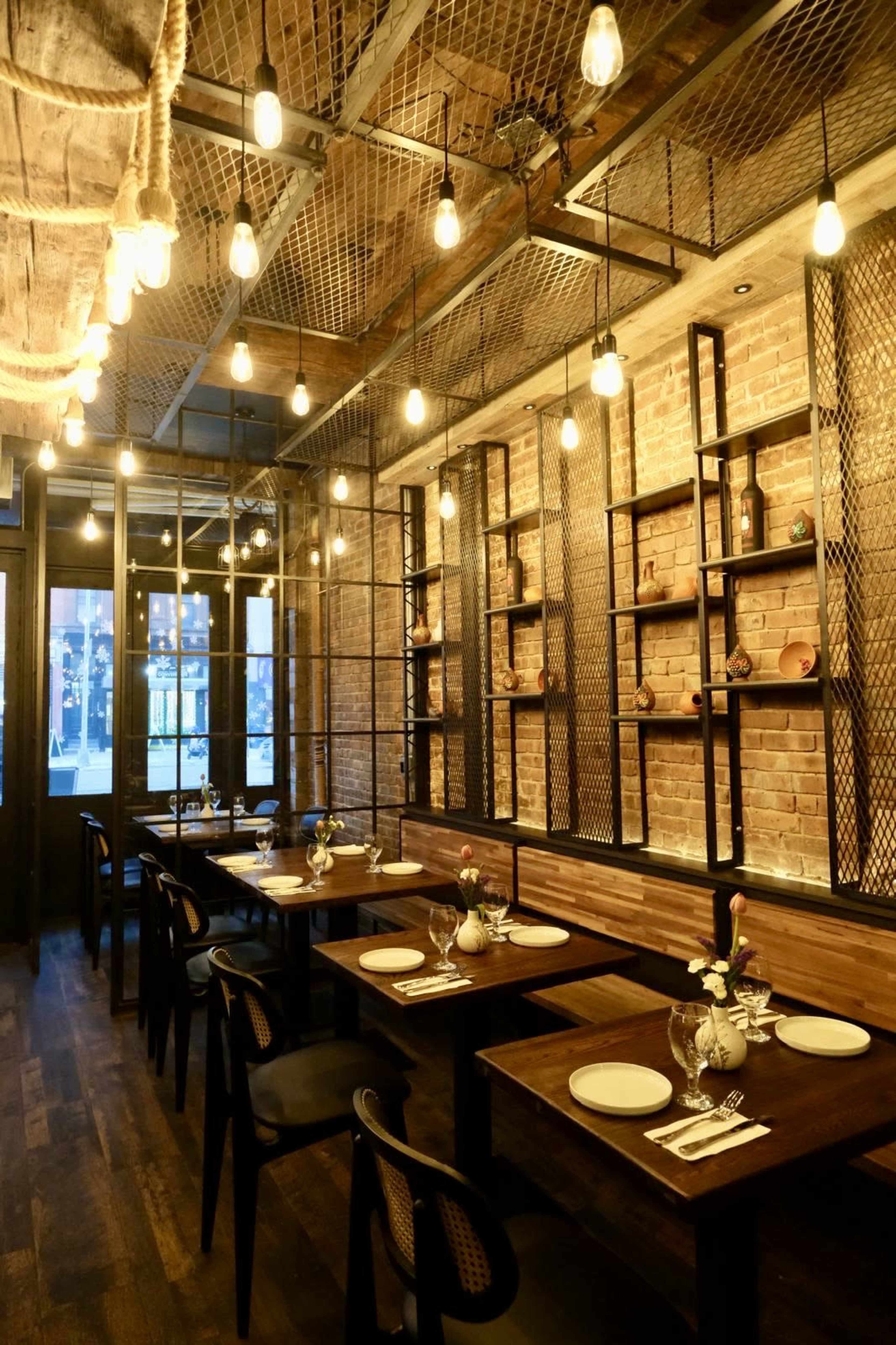 The image shows a modern restaurant interior with wooden tables set for dining, industrial-style lighting fixtures, and exposed brick walls.